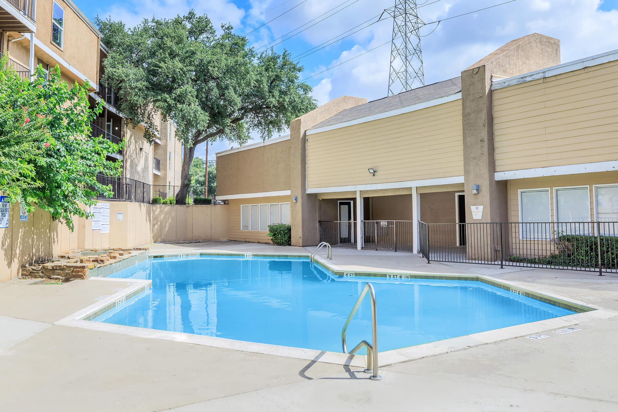 Swimming pool surrounded by beige apartment buildings. Lush green trees and a clear blue sky in the background. A fence encloses the pool area, and there are power lines visible in the distance. The pool has a shallow entry and steps for easy access.
