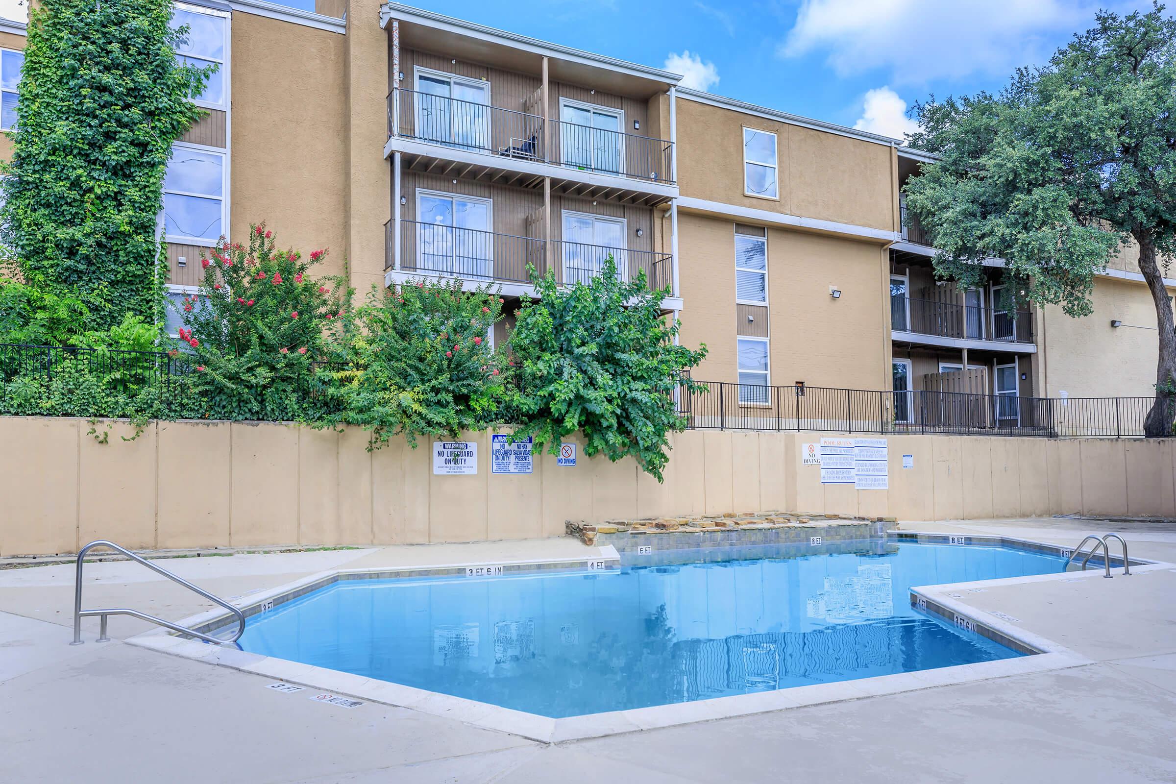 A clear blue swimming pool surrounded by a concrete deck, with a beige apartment building in the background featuring balconies. Lush greenery, including trees and bushes, adds a natural touch to the area. The pool area appears well-maintained and inviting, perfect for relaxation.