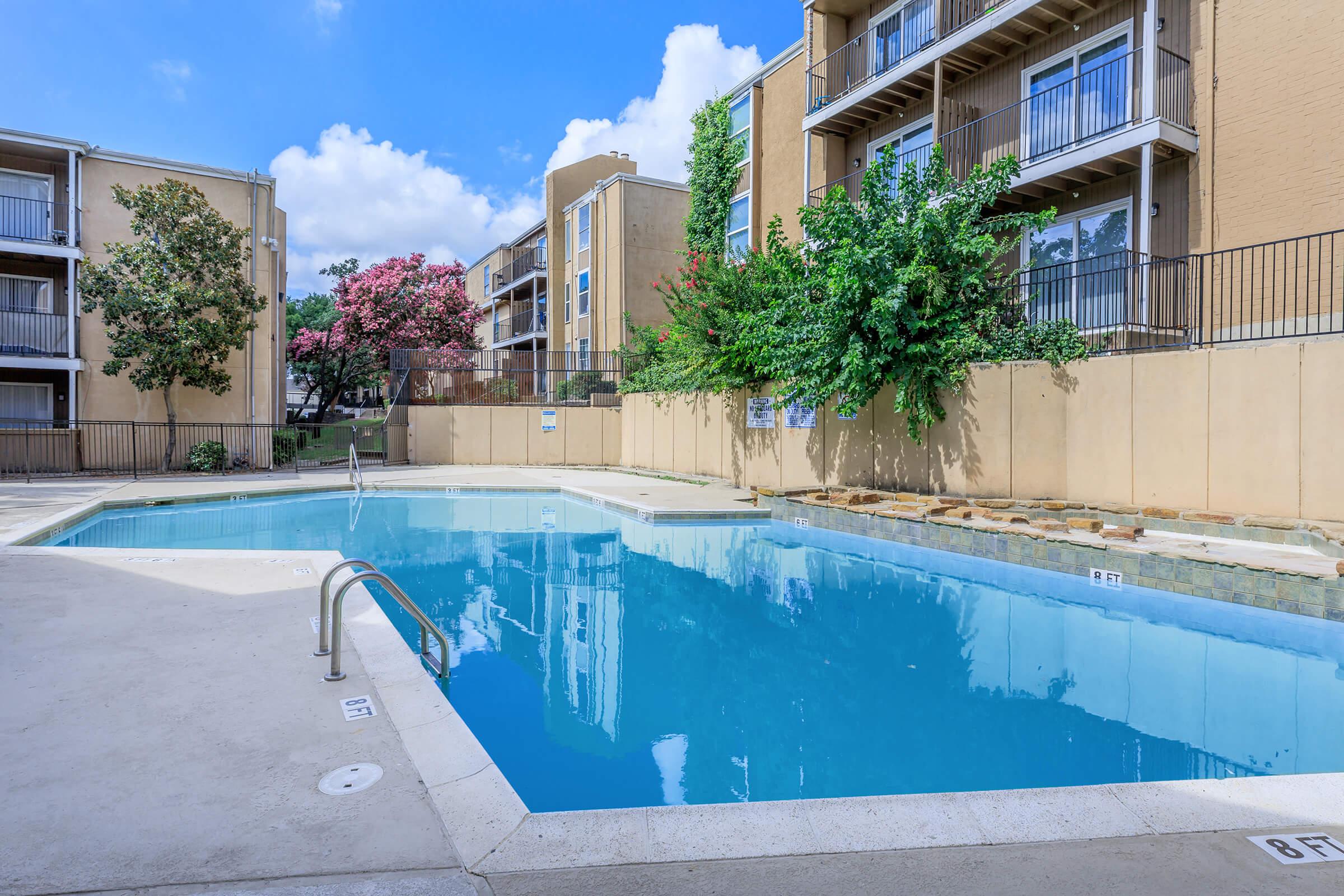 Swimming pool surrounded by apartment buildings, featuring clear blue water. Lush green trees and flowering plants are visible nearby, under a bright blue sky with fluffy white clouds. The area is well-maintained, inviting relaxation and leisure activities.