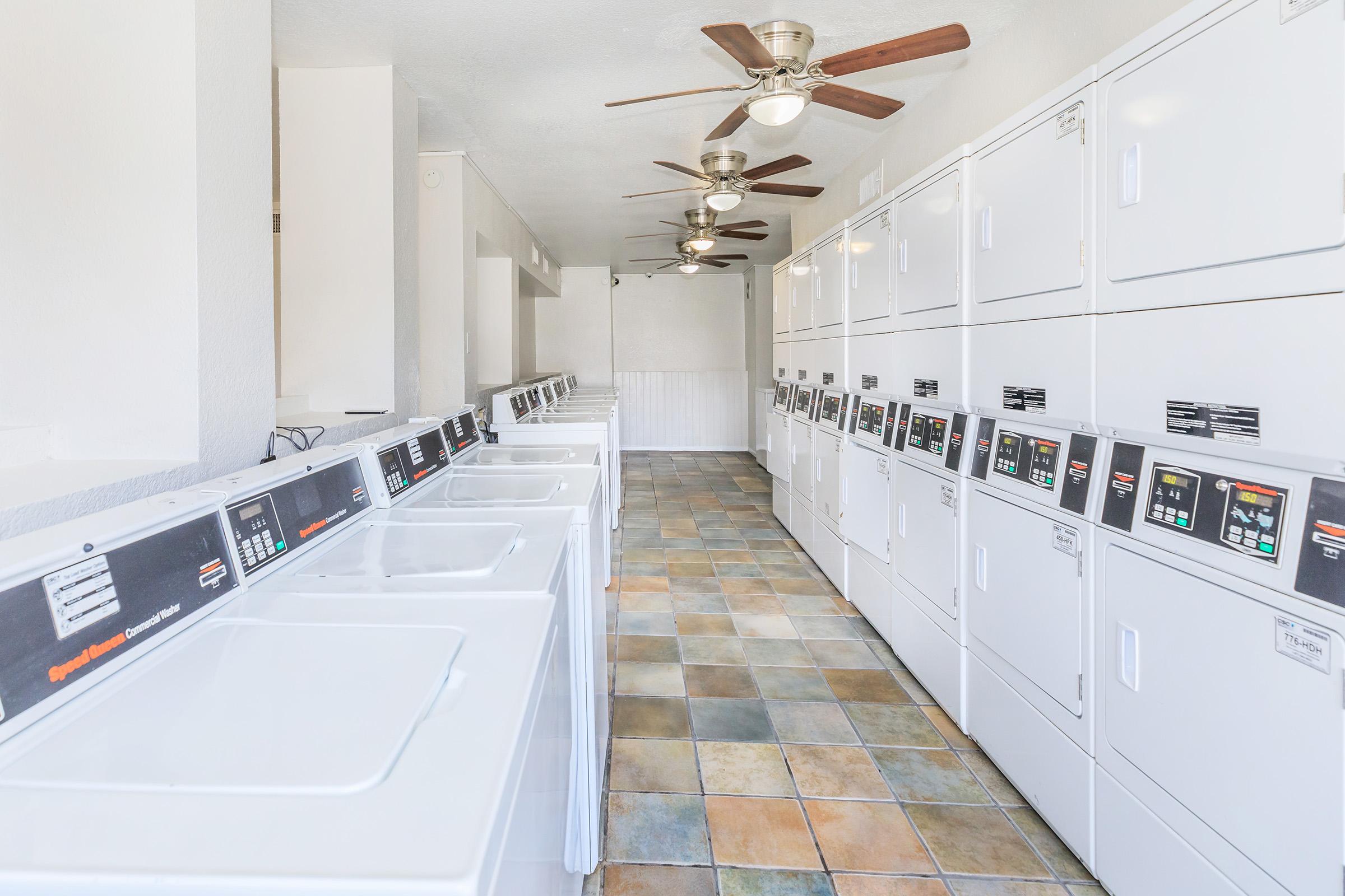 A clean and well-lit laundromat interior featuring rows of white washing machines and dryers. The space includes ceiling fans and colorful tiled flooring, creating a welcoming atmosphere. Machines are equipped with digital displays for ease of use.