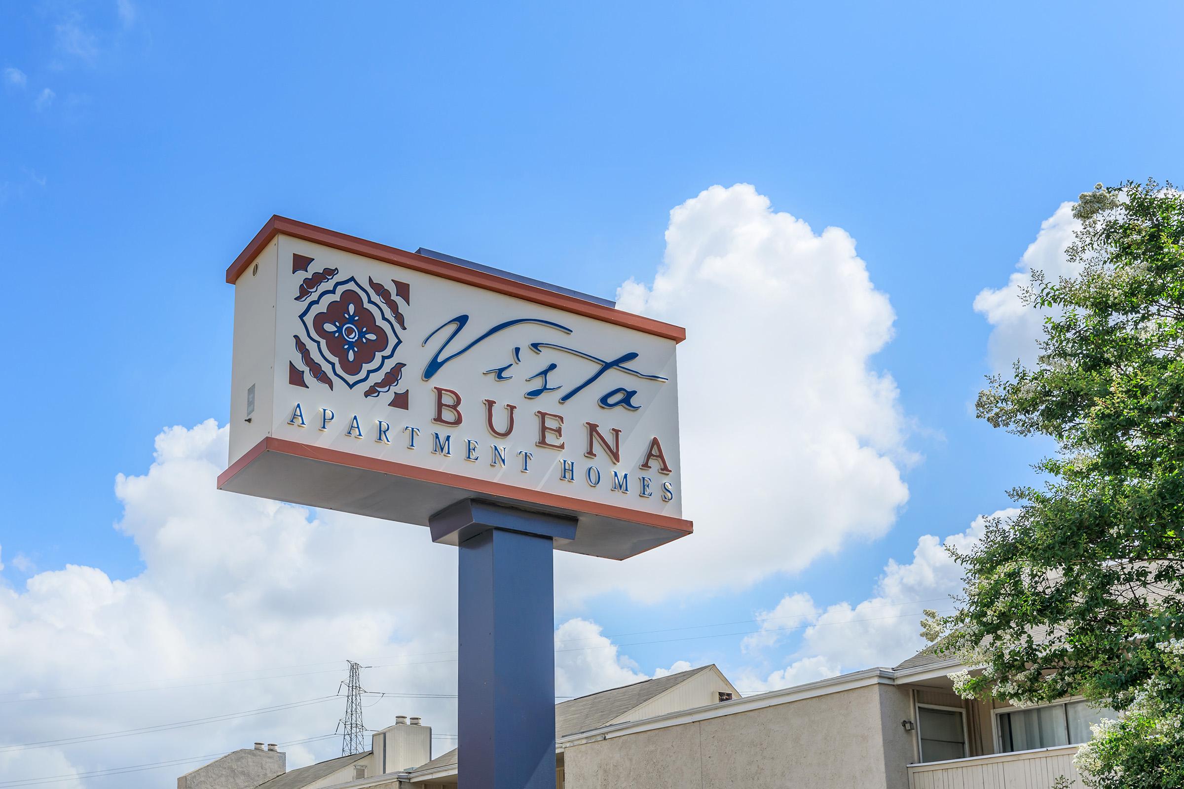 Sign for "Vista Buena Apartment Homes" against a blue sky with white clouds. The sign features a decorative emblem and is mounted on a post, indicating the name of the apartment complex.