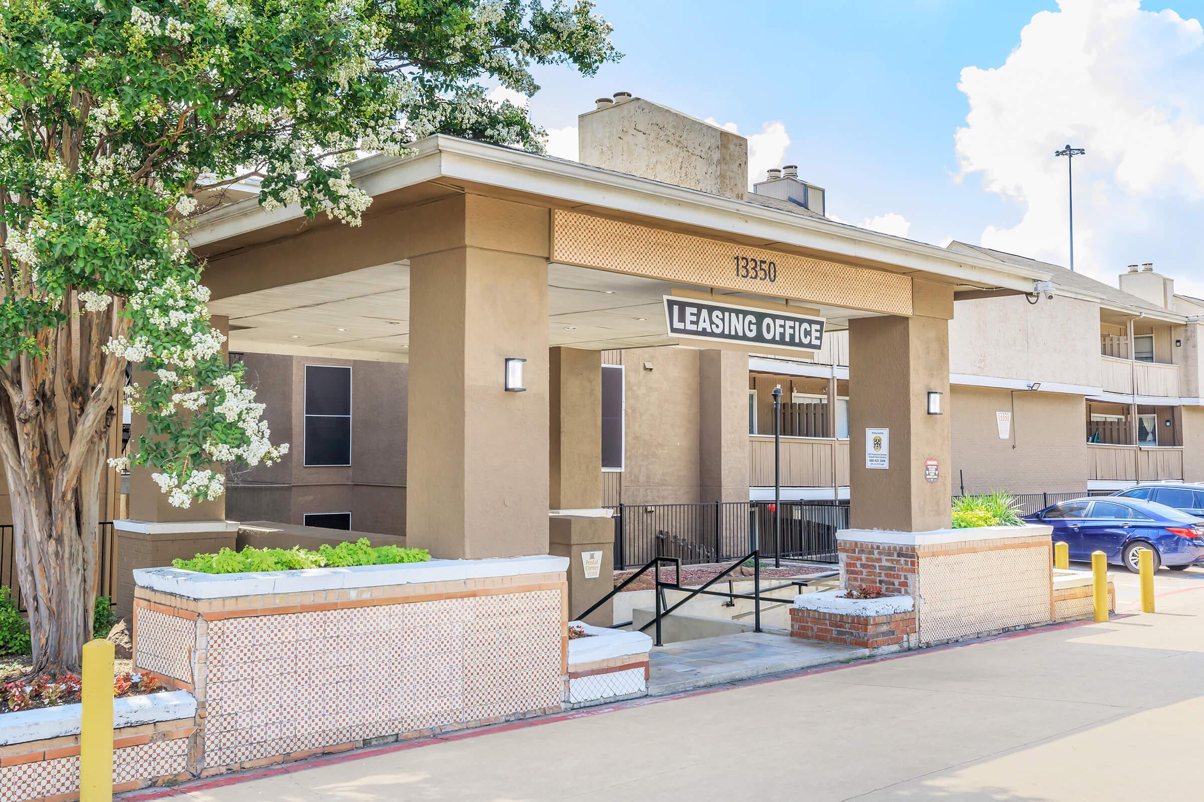 Leasing office entrance with a prominent sign, surrounded by greenery and decorative landscaping. The building features a modern design with a neutral color palette. There is a set of stairs leading to the main door and vehicles parked nearby. Bright blue sky and fluffy clouds are visible in the background.
