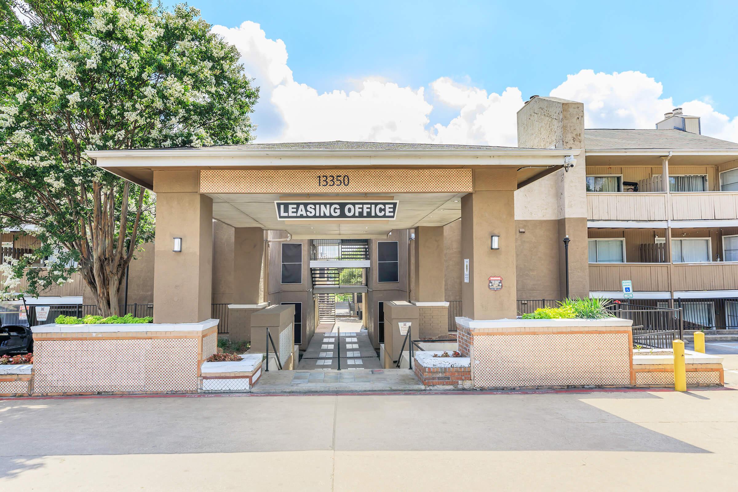 Leasing office entrance featuring a modern architectural design with a sign that reads "Leasing Office." The building is surrounded by greenery and has steps leading to the entrance. Blue sky and clouds are visible in the background, creating a bright and inviting atmosphere.