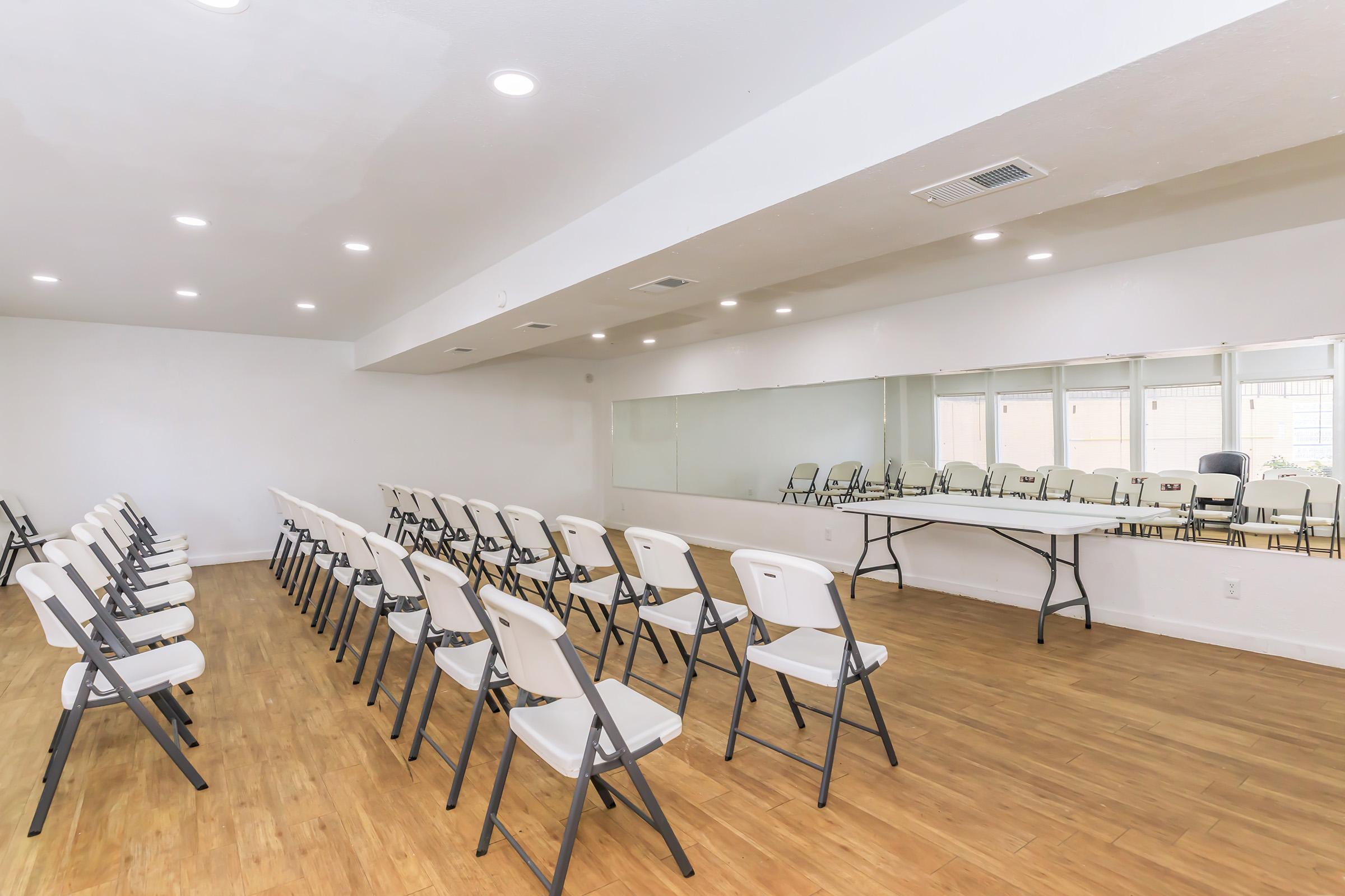 A spacious room with a wooden floor, featuring rows of white folding chairs facing a long table. A large mirror reflects the seating area, and there are windows allowing natural light to fill the space. The walls are painted white, creating a bright and clean atmosphere suitable for events or meetings.