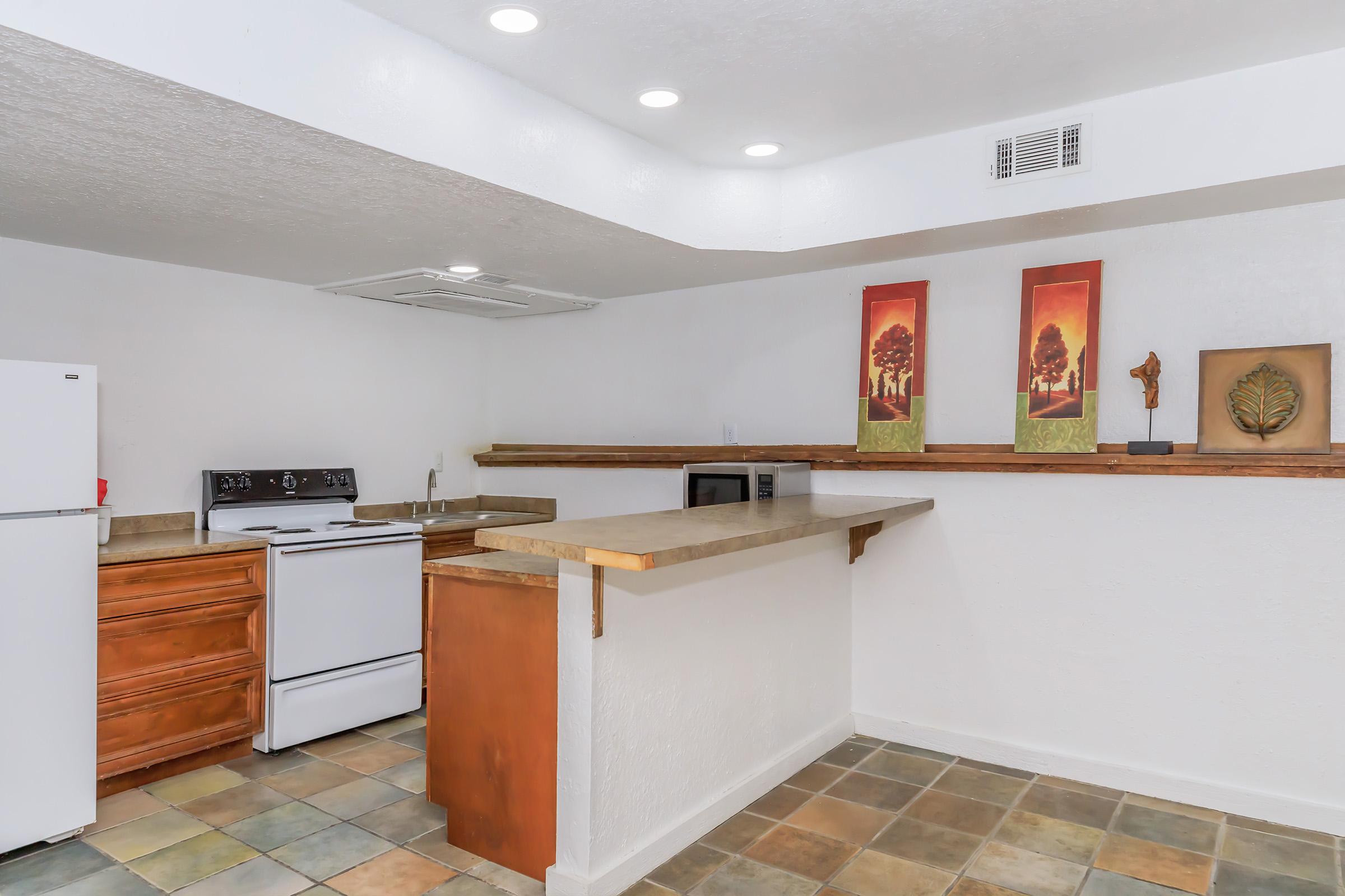 Modern kitchen with a white refrigerator, stove, and microwave. The countertops are stone, with wooden cabinetry on one side. The floor features multicolored tiles, and there are two framed artworks on the wall depicting trees. Bright lighting enhances the spacious and clean design.
