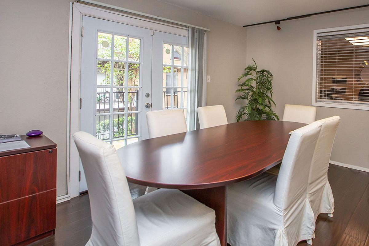 A modern dining area featuring a large oval wooden table surrounded by six white upholstered chairs. In the background, there are glass double doors leading to a balcony, a potted plant, and a window with blinds. The room has a neutral color scheme and wooden flooring.
