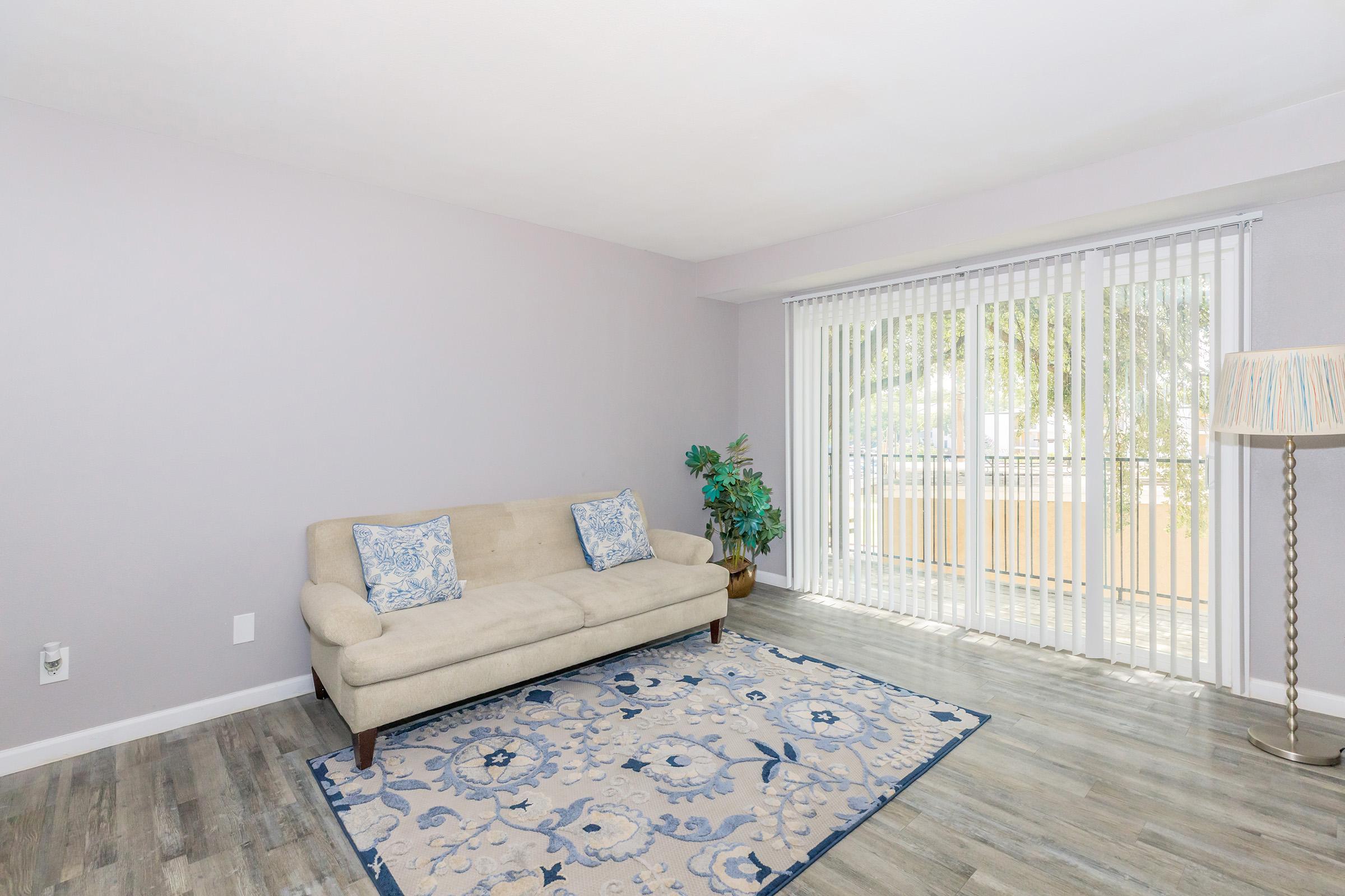 A light and airy living room featuring a beige sofa with blue throw pillows, a decorative area rug with floral patterns, a potted plant in the corner, and large sliding glass doors with vertical blinds letting in natural light. The walls are painted in a soft lavender hue, enhancing the tranquil atmosphere.