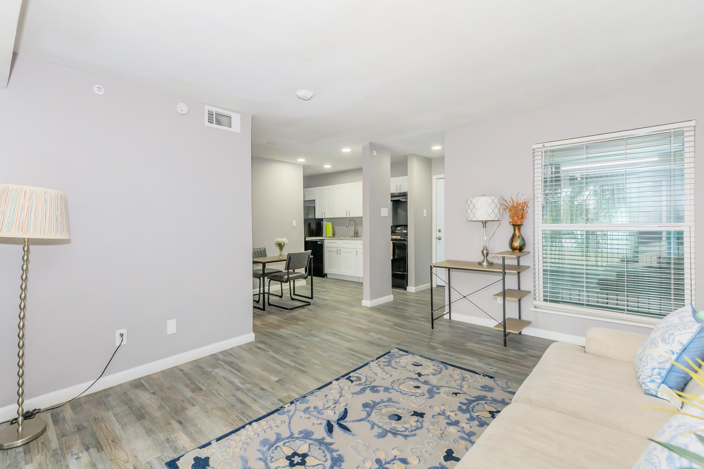A modern living space featuring light gray walls, a beige couch, and a decorative rug. A small dining area with a table and chairs is adjacent to a kitchen, which is partially visible in the background. Natural light streams through a window, creating a bright and inviting atmosphere.