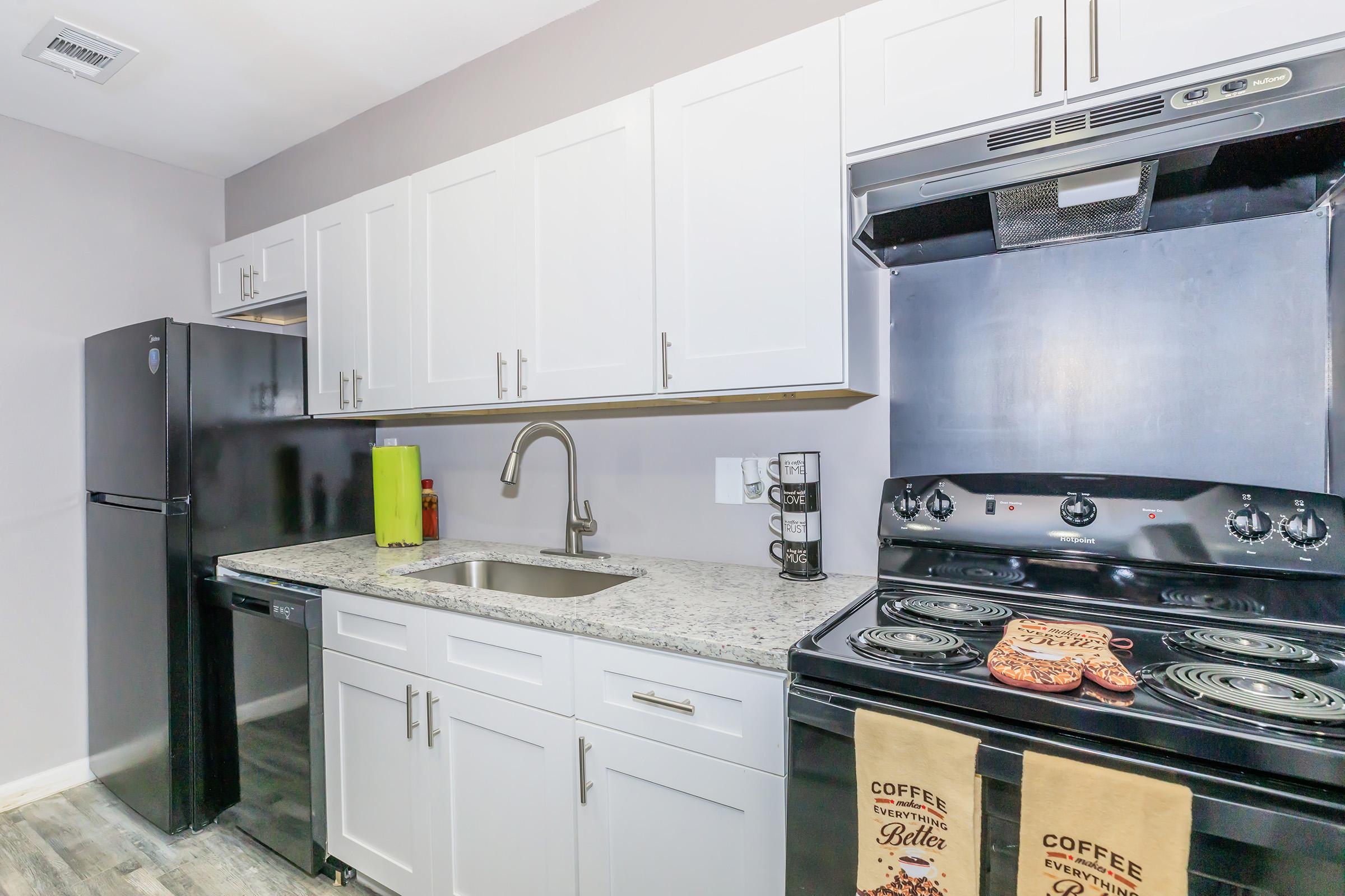 Modern kitchen featuring white cabinetry, a stainless steel sink with a pull-down faucet, a black refrigerator, and an electric stove with an oven. The countertop is made of granite, and two decorative towels with coffee-themed text hang from the stove. The walls are painted in a light gray color.
