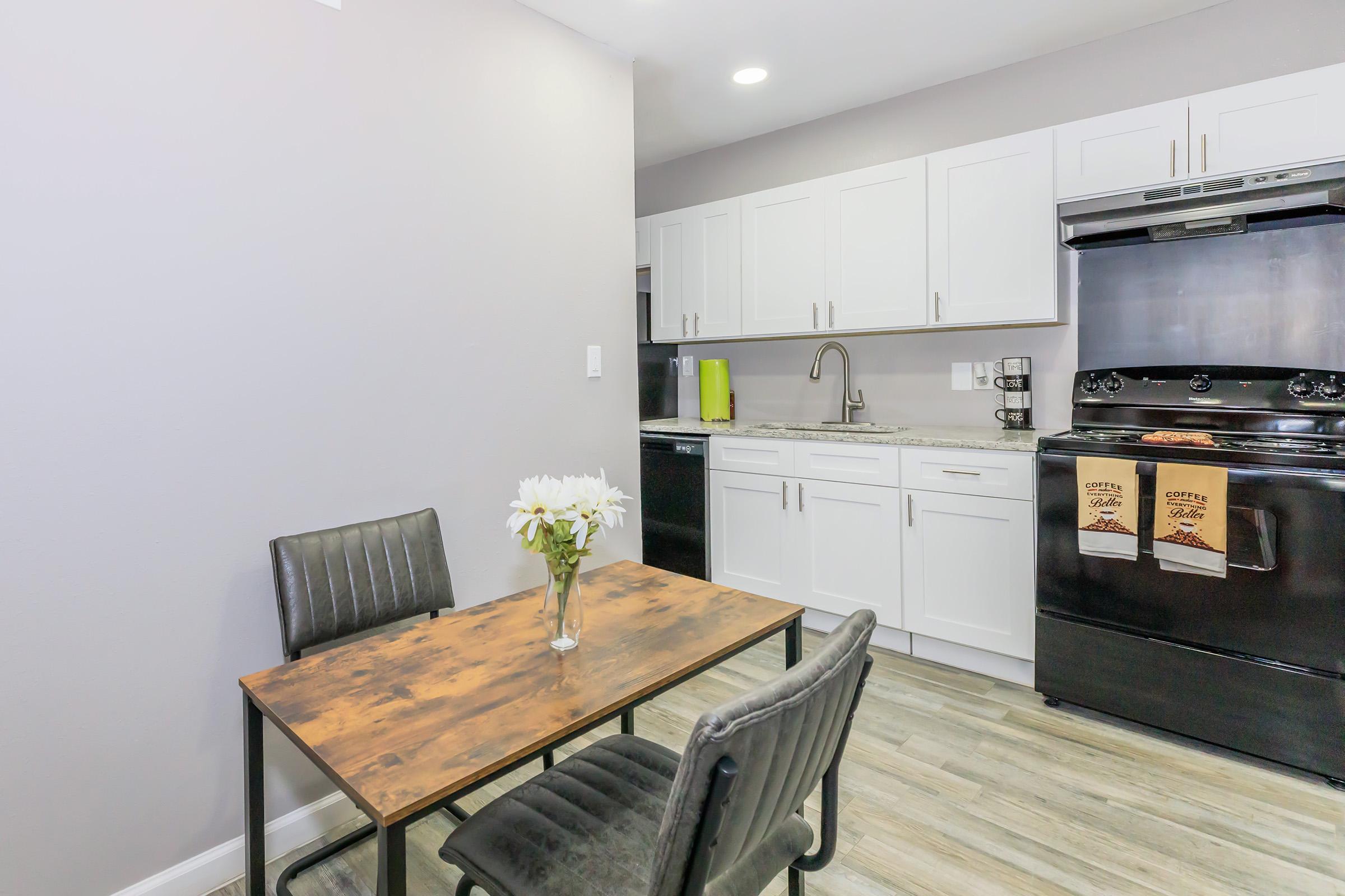 A modern kitchen with white cabinets and black appliances. A small wooden dining table with two black chairs sits in the foreground, featuring a vase of white flowers. The kitchen has a sleek design with a light gray wall and wood-look flooring.