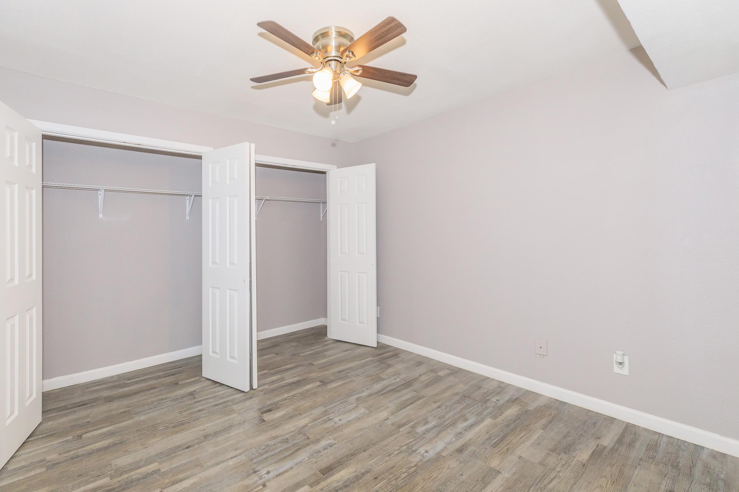 A well-lit room with a ceiling fan and light fixture. The walls are painted in a soft lavender shade. Two white double doors open to spacious closets. The floor features a light wood laminate, enhancing the room's modern aesthetic and open feeling.