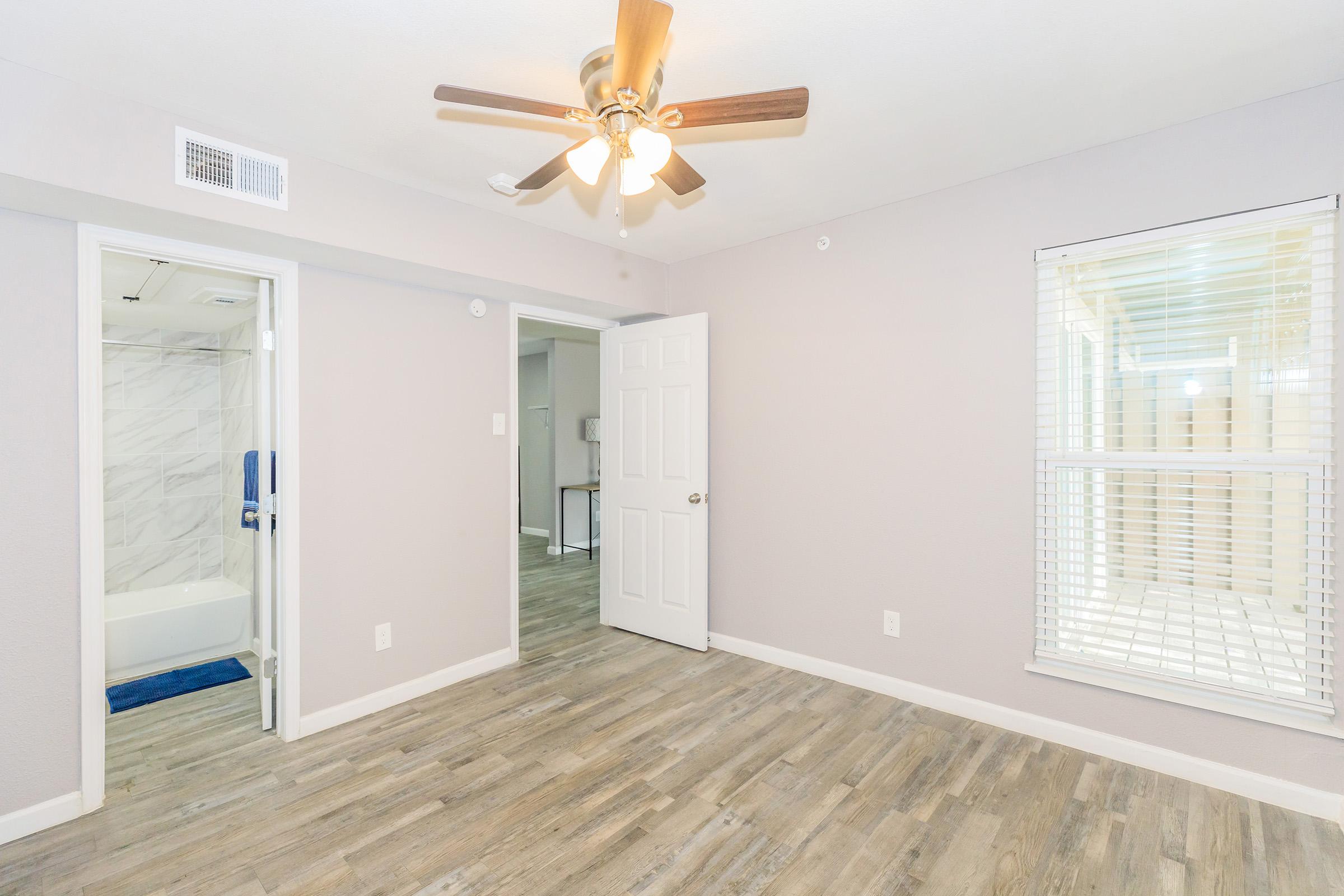 A spacious room featuring light-colored walls, a ceiling fan, and wooden flooring. A doorway on the left leads to a bathroom with marble tiles and a bathtub. Another door opens to a small hallway, while a large window allows natural light to fill the space.