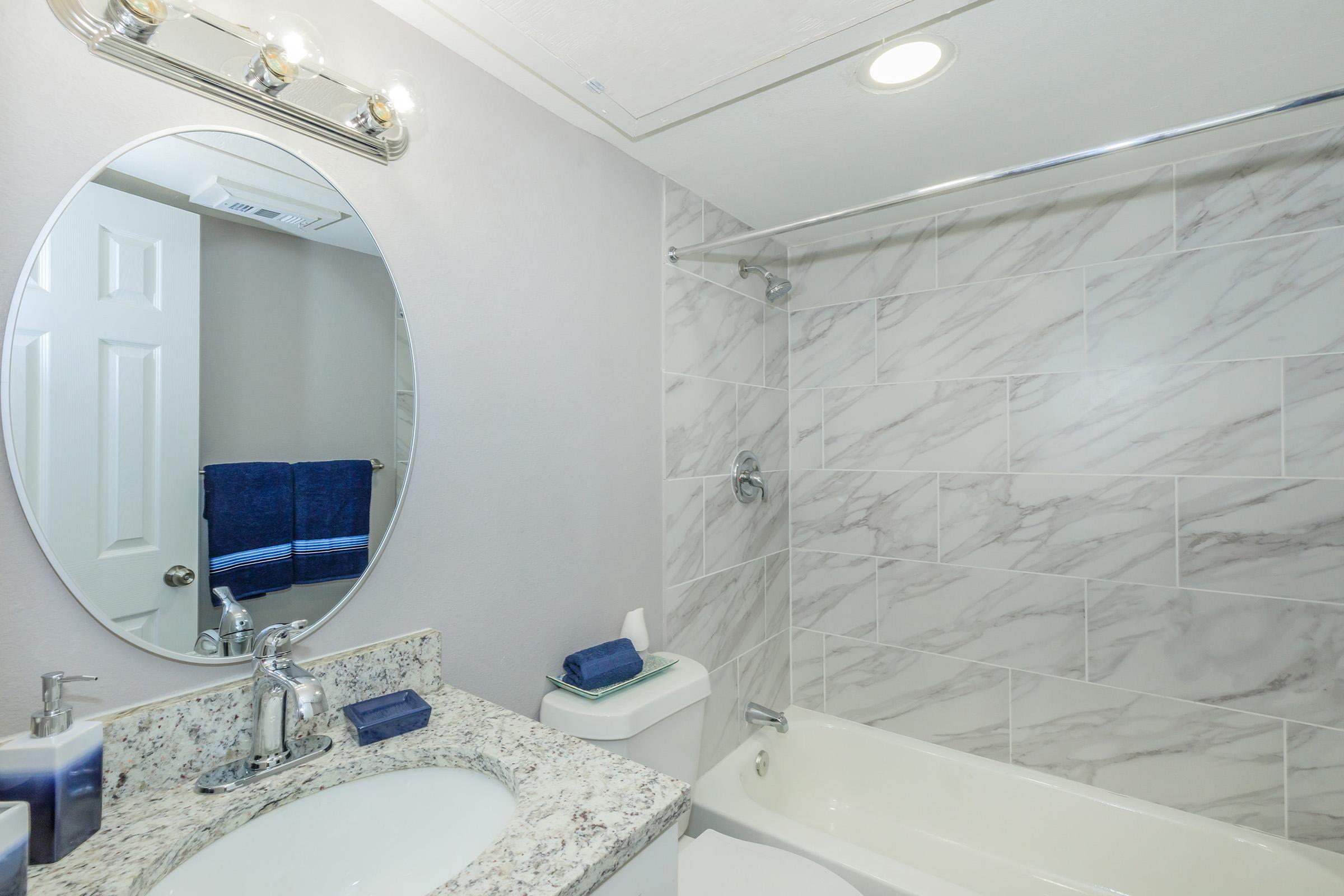 A modern bathroom featuring a circular mirror above a granite countertop with a sink. The walls are covered in light marble tiles, and there is a bathtub with a shower rod. A neatly arranged set of blue towels is visible in the background, along with toiletries on the counter. The overall aesthetic is clean and contemporary.
