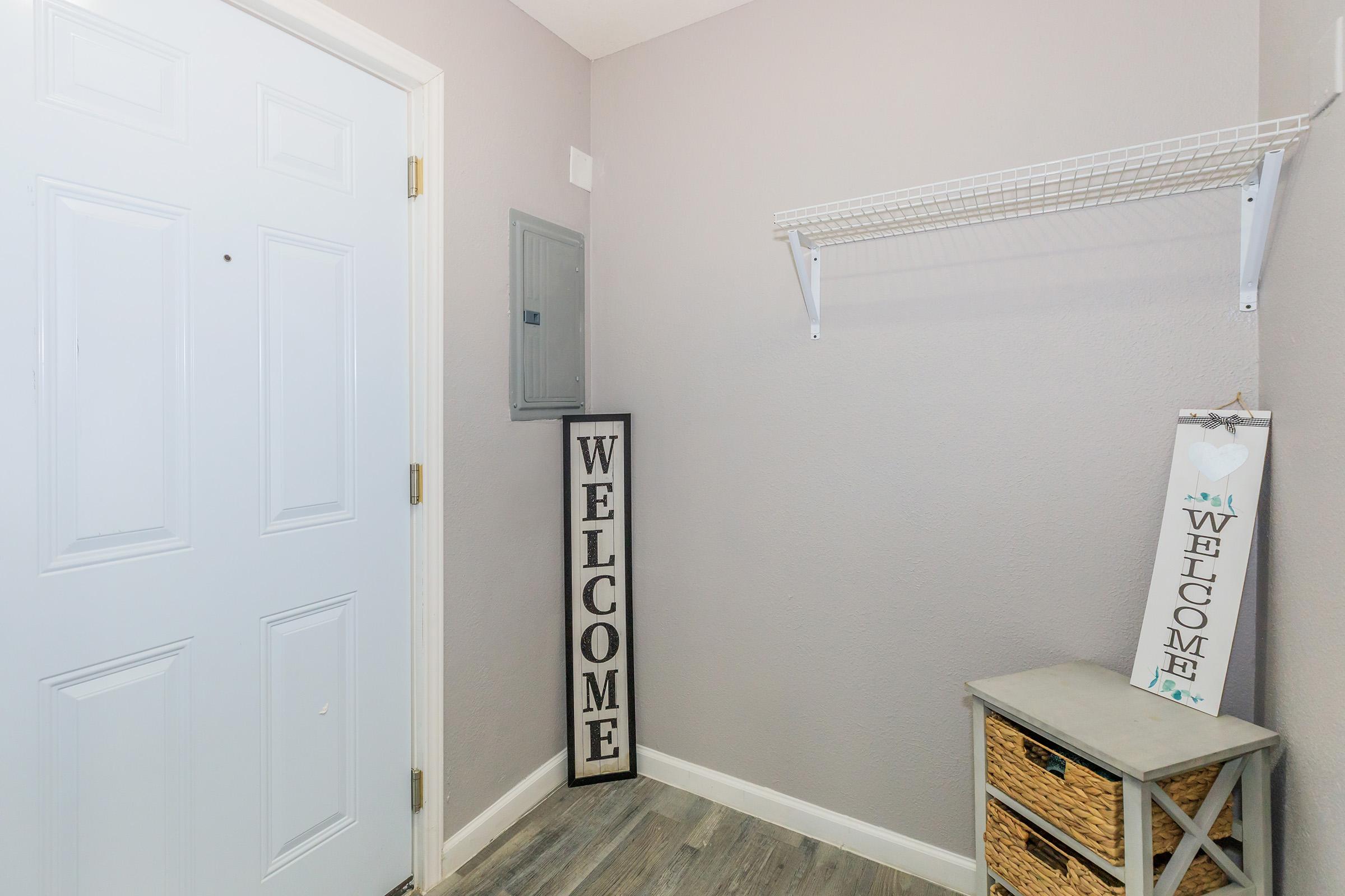 A cozy entryway featuring a light gray wall, a white door, and two welcome signs—one tall and vertical, the other shorter. There's a wall-mounted shelf and a small table with a woven basket underneath, creating an inviting atmosphere for guests.