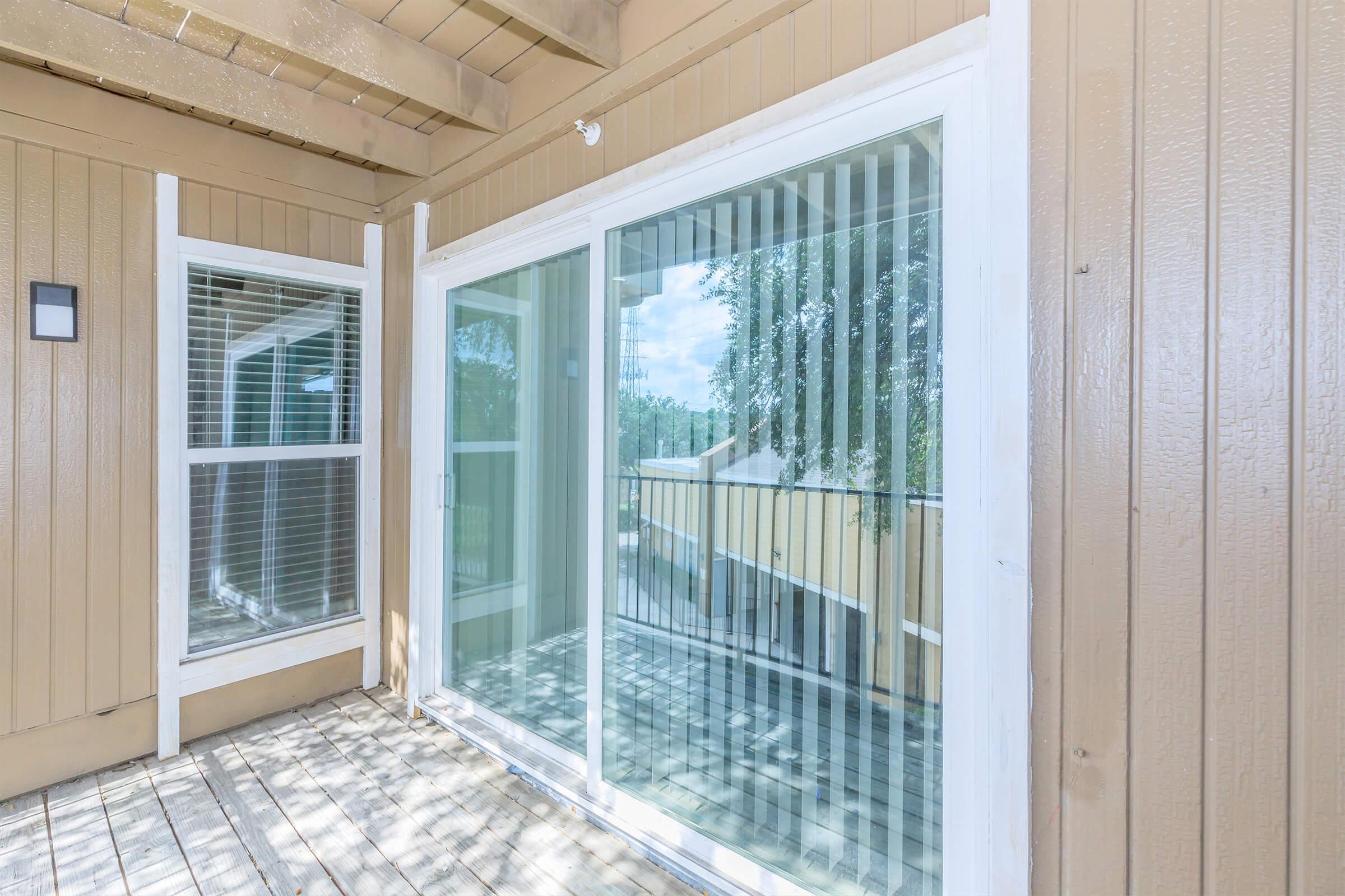 A view of a wooden deck area featuring large sliding glass doors with vertical blinds. The exterior walls are painted in a warm brown color, and there are windows on the left side allowing natural light to enter the space. The surrounding area includes greenery visible through the glass.