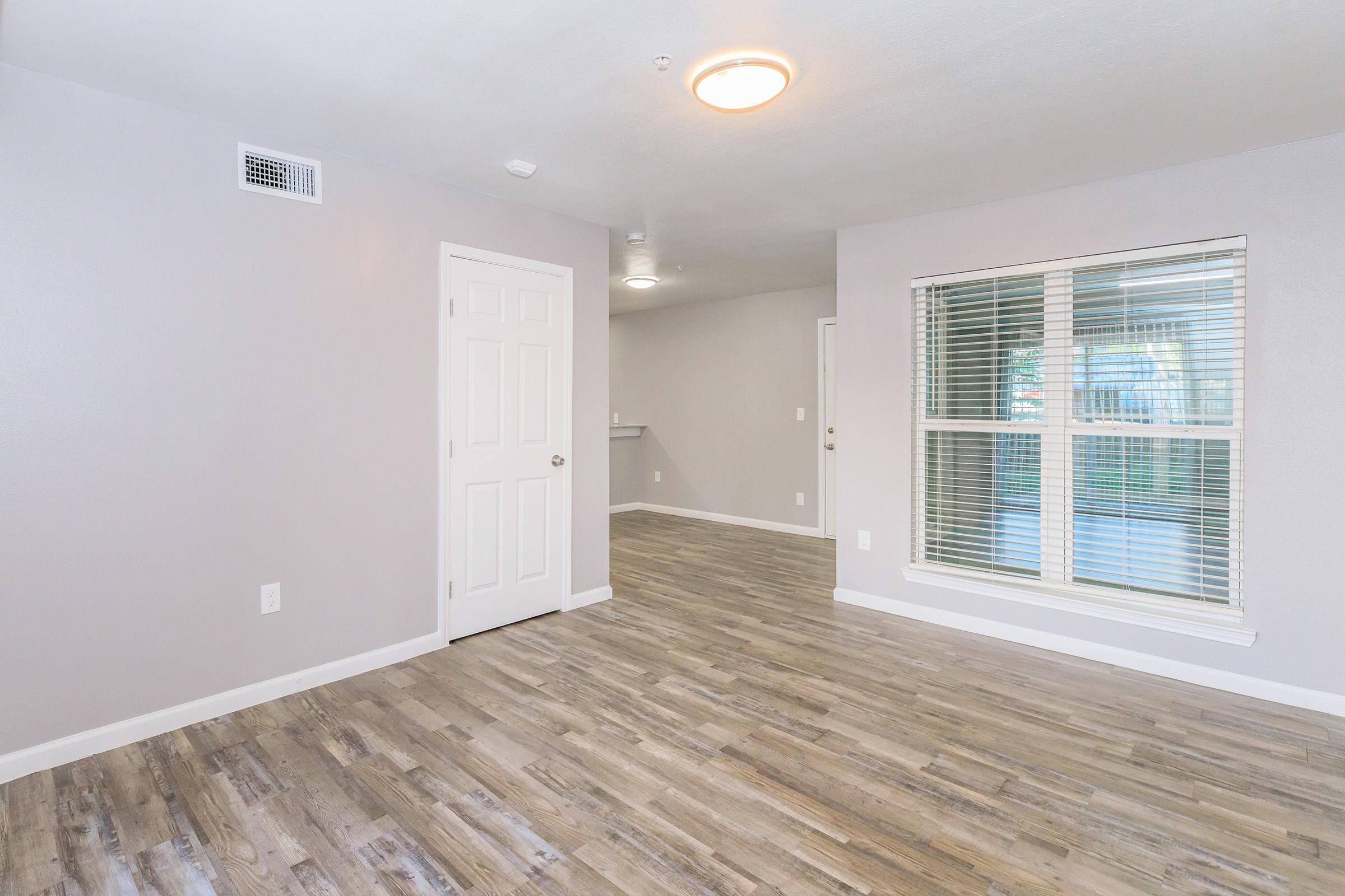 A spacious, bright living area with light gray walls and a modern wood-like floor. A large window allows natural light to fill the room, while a closed white door is visible on the left. The area is well-maintained and inviting, suitable for various furniture arrangements.