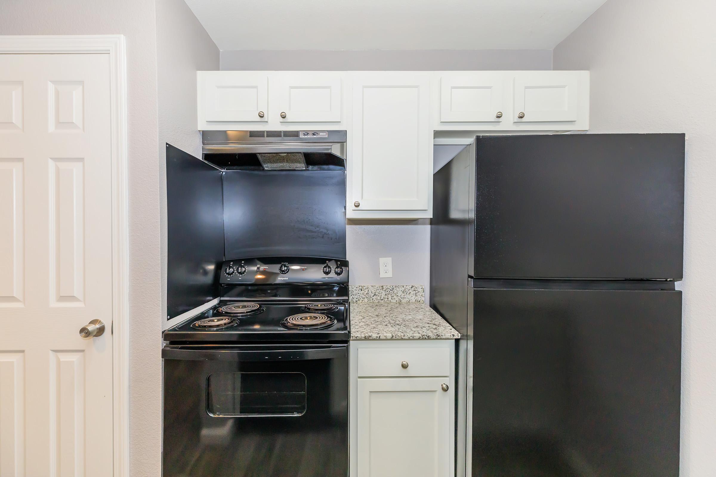 A compact kitchen featuring a black stove and refrigerator, with white cabinets above a granite countertop. The space has light-colored walls and a closed white door, giving it a clean and modern appearance.