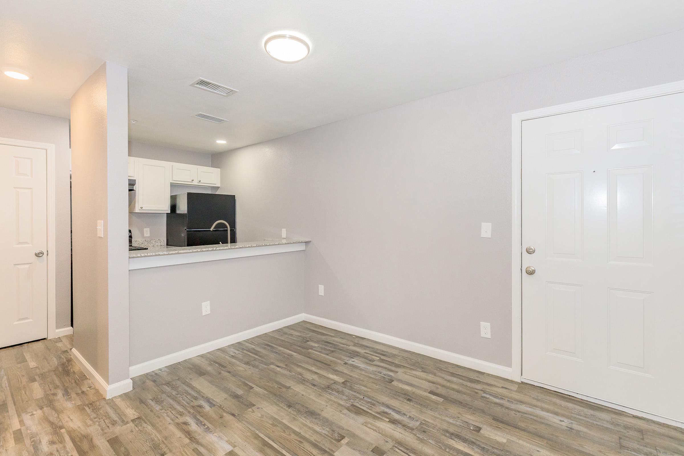 Interior view of a modern, empty apartment showcasing light gray walls and wood-like flooring. The space features a small kitchen area with a countertop, a white door leading outside, and a door on the left, possibly leading to a closet or another room. Soft ceiling lighting illuminates the room.