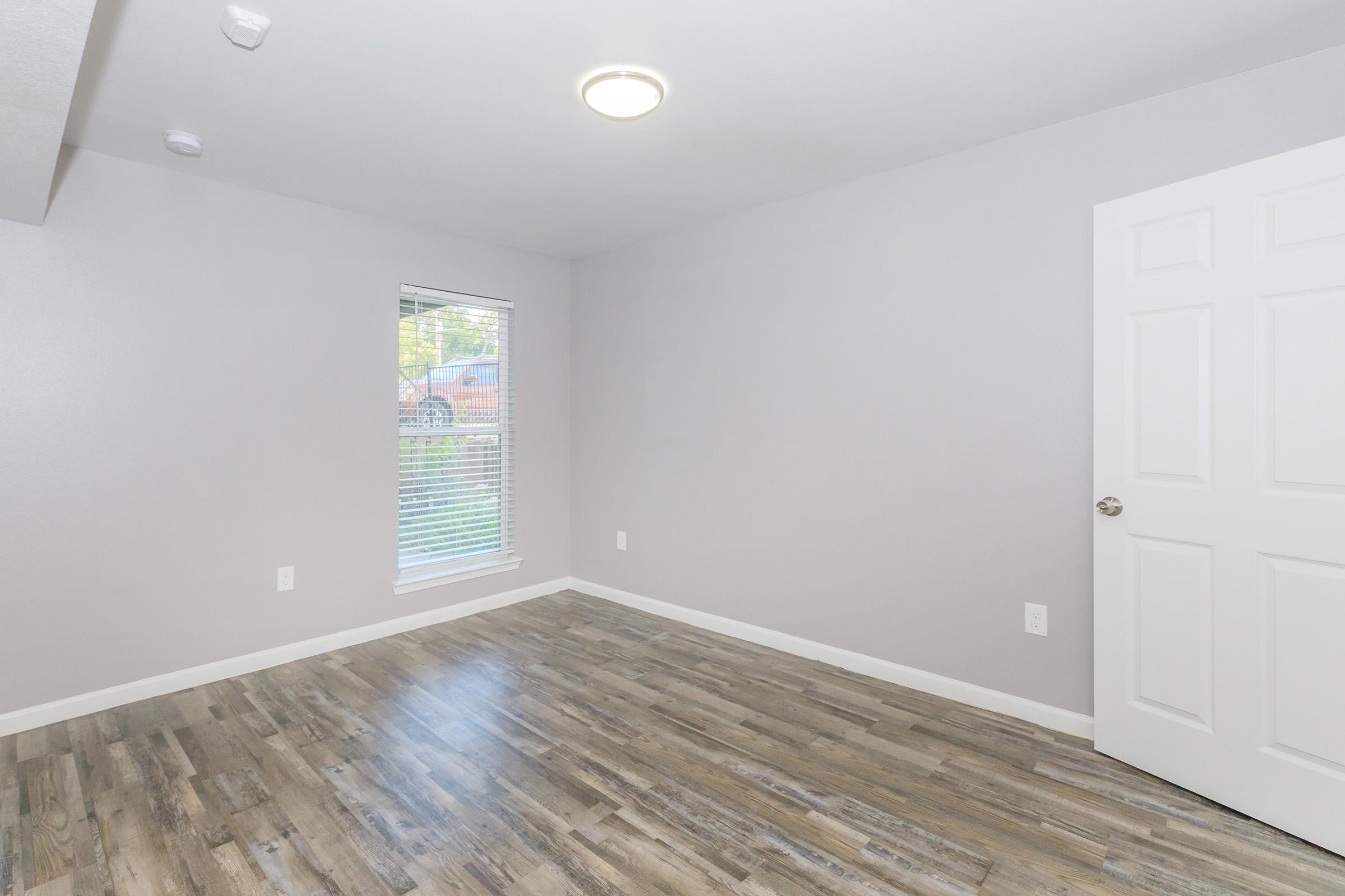 Empty room with light gray walls, laminate flooring, and a single window with blinds allowing natural light. A closed white door is on the right side, and ceiling lights provide illumination. The space is minimalist and ready for furnishing.