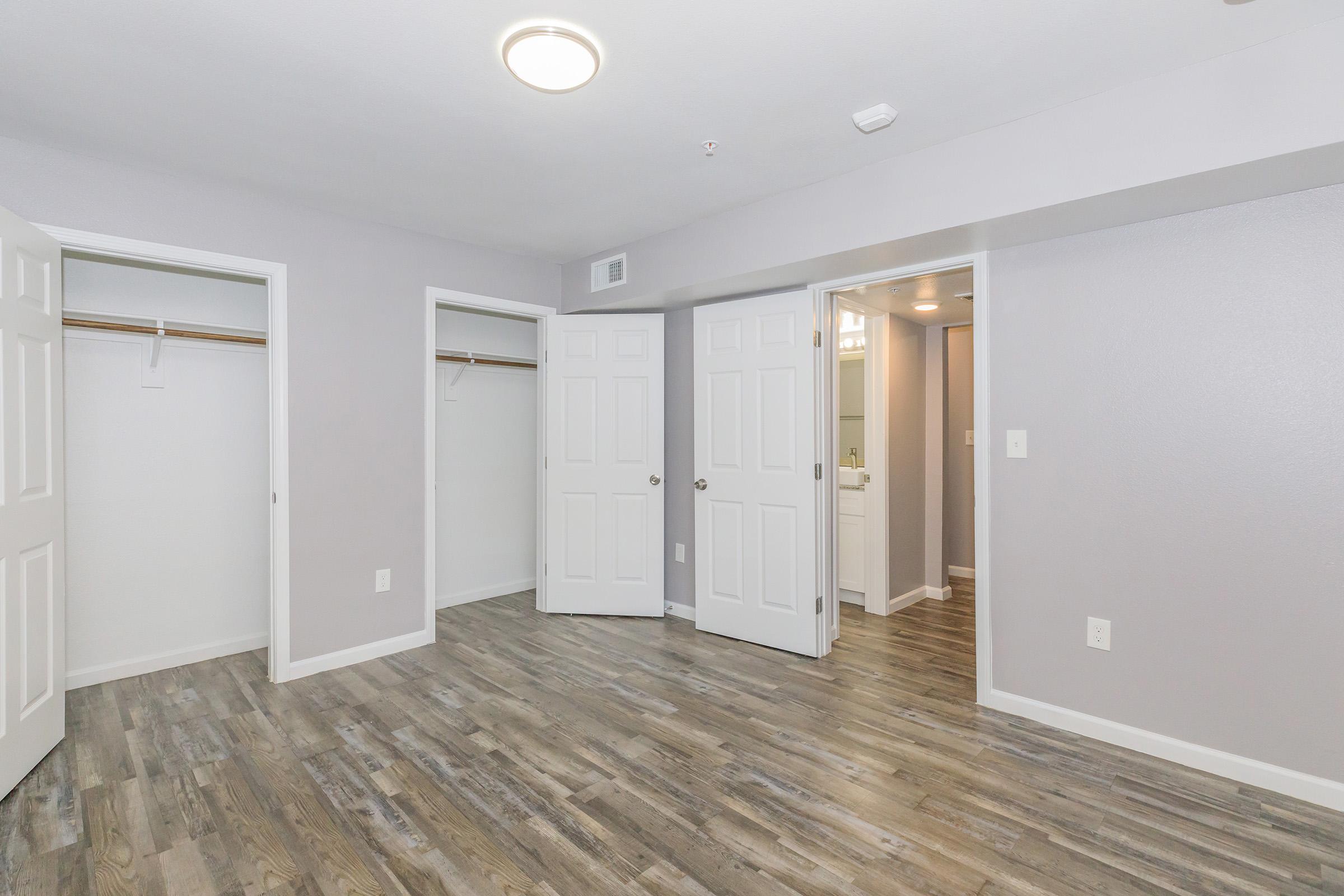 A spacious room featuring light gray walls, two open closet doors on the left, and a doorway to a bathroom on the right. The floor is made of wood-like laminate, and there is a ceiling light fixture. The overall ambiance is modern and well-lit.