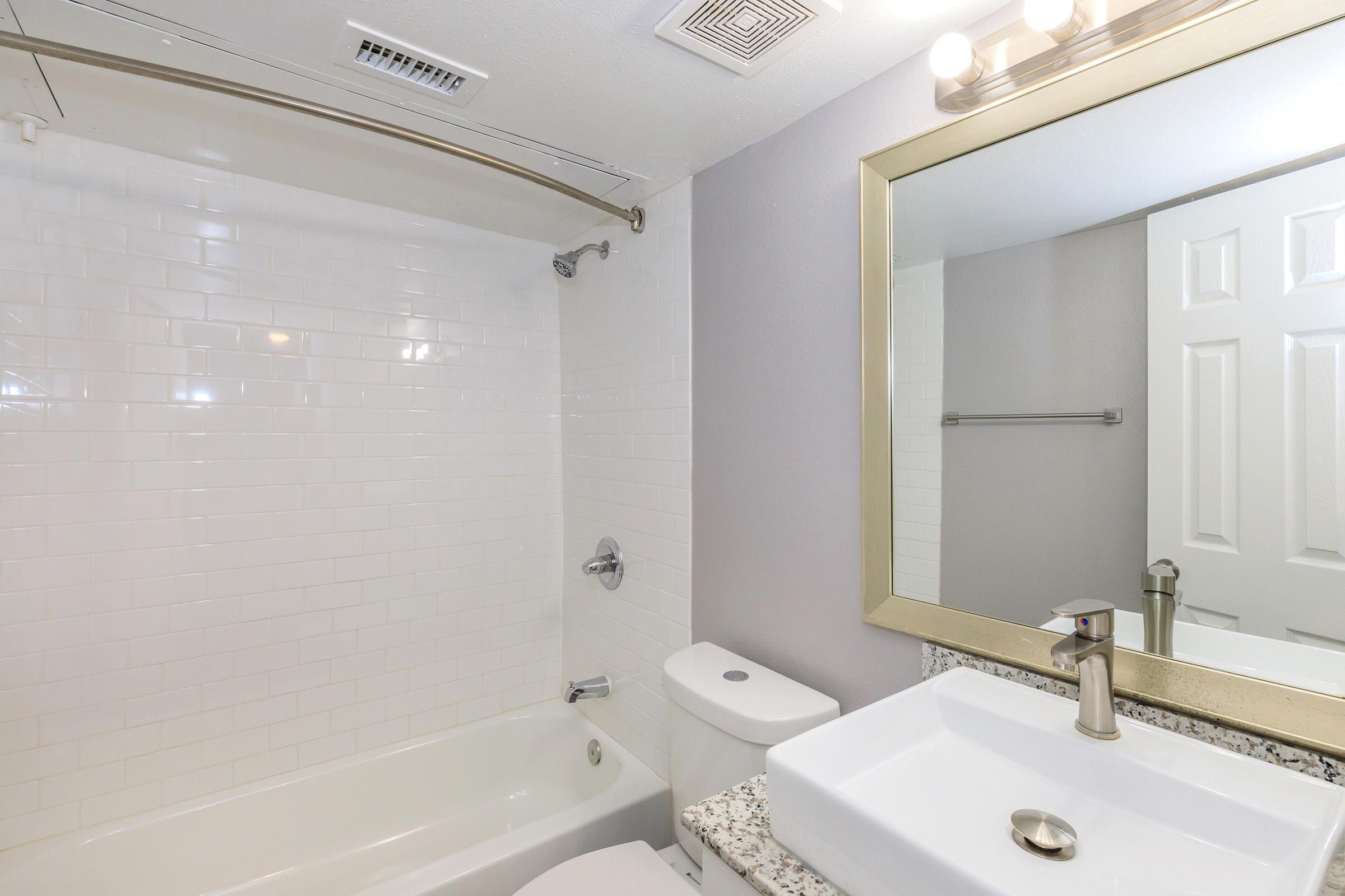 Modern bathroom featuring a bathtub with a shower, white tiled walls, a granite countertop sink, and a large mirror with decorative framing. The space is well-lit with a ceiling light fixture, and includes basic bathroom fixtures for functionality.