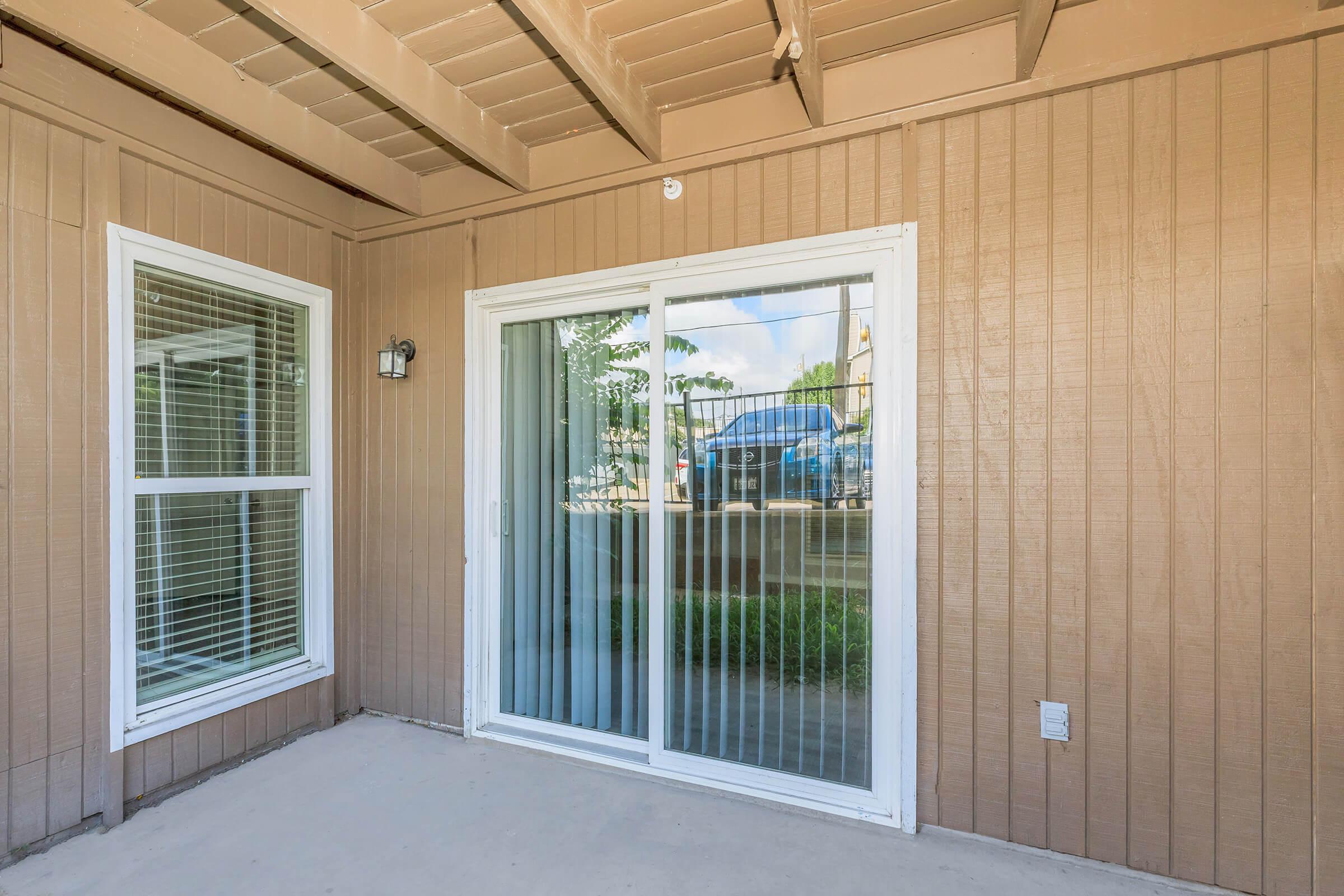 Sliding glass door leading to a patio area, with vertical blinds partially closed. The exterior wall is made of horizontal wood planks, and there are windows on either side of the door. Natural light is visible, enhancing the outdoor view.