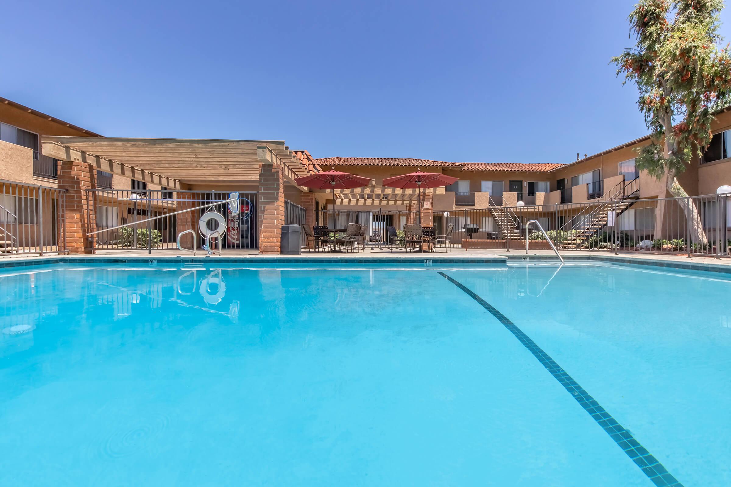 A clear blue swimming pool is shown in the foreground, with lounge chairs and red umbrellas arranged around it. In the background, there are two-story buildings with balconies and a wooden pergola, under a bright blue sky. The scene conveys a relaxing, inviting atmosphere.