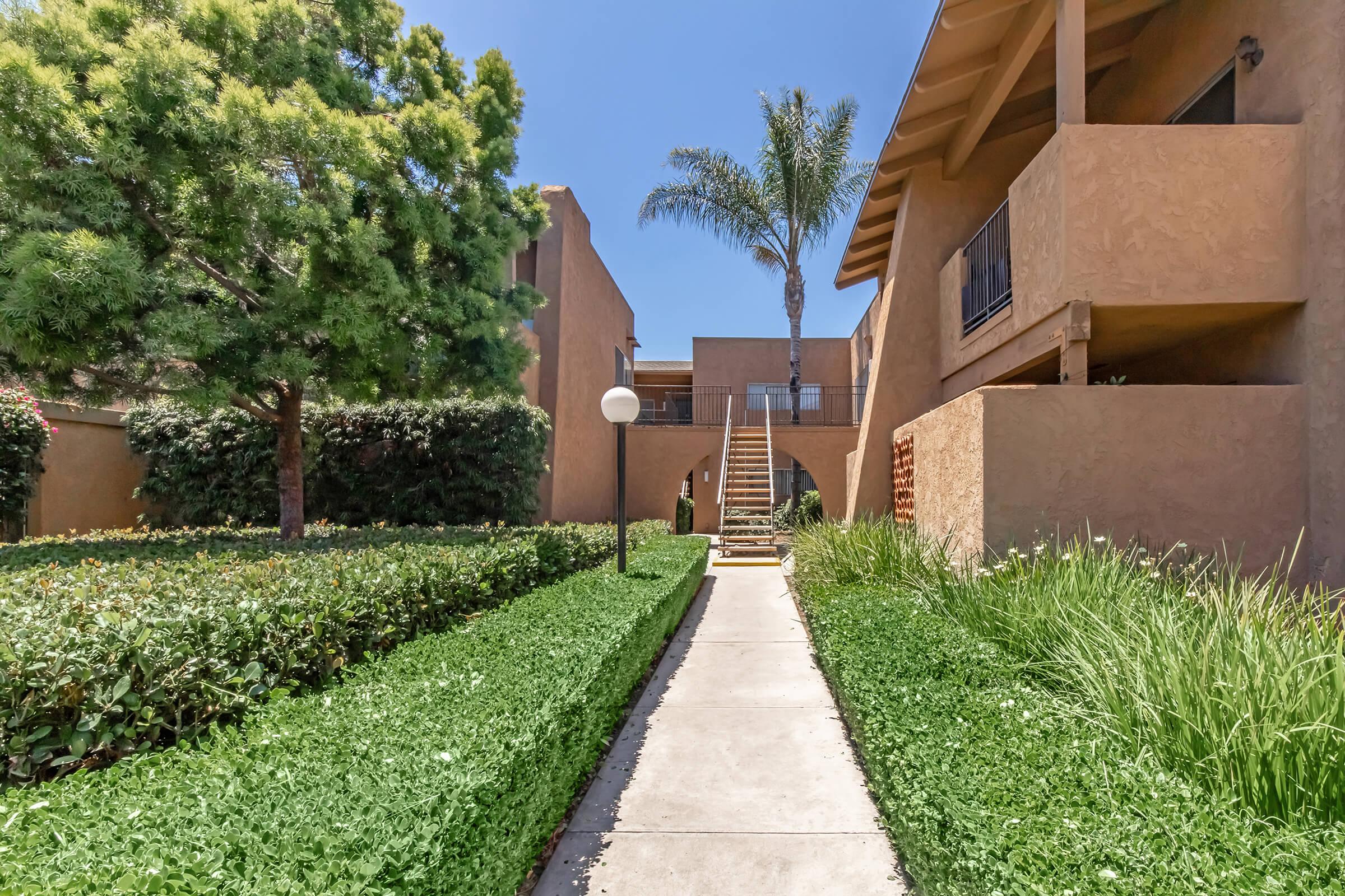 Pathway lined with green shrubs and grass leading towards a set of apartment buildings. There are trees and a palm tree in the background, with a light fixture along the pathway. The buildings feature a warm, tan stucco exterior under a clear blue sky.