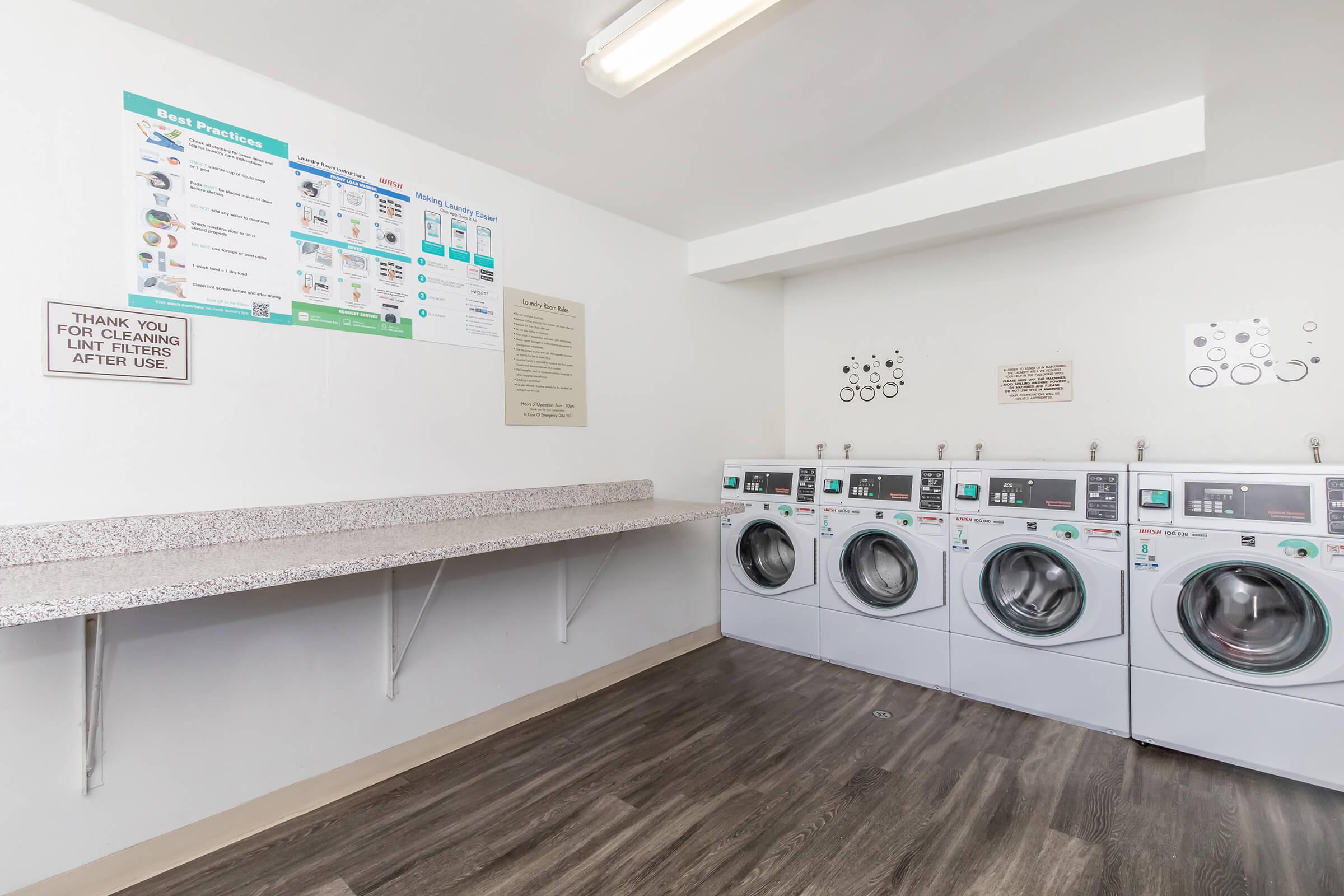 A clean laundry room featuring three front-loading washing machines and a countertop for folding. Instructions and guidelines are posted on the wall, and there is ample space for laundry activities in a well-lit environment.