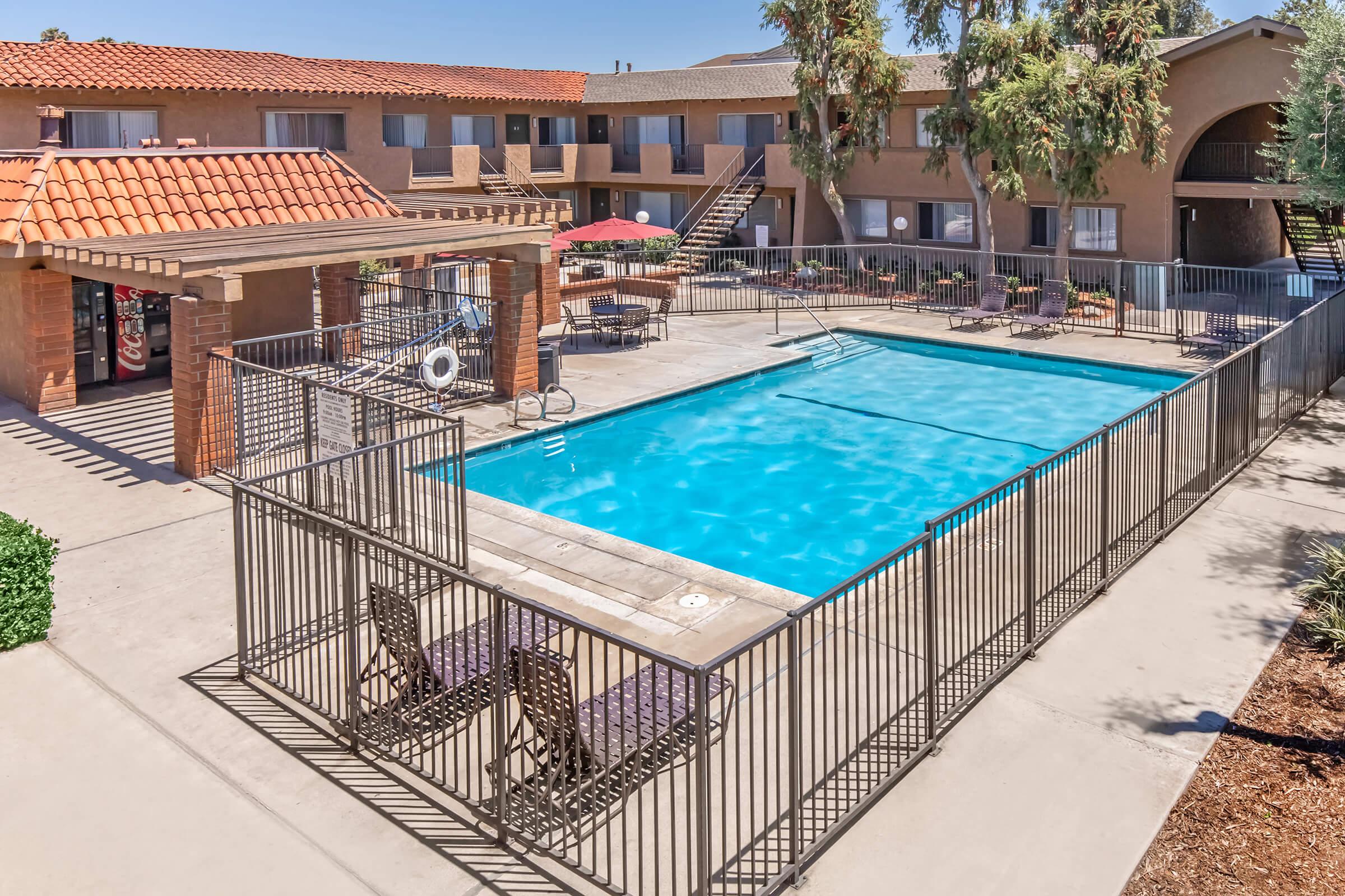 An outdoor swimming pool surrounded by a gated enclosure, with lounge chairs on the pool deck. Nearby, there are shaded areas with umbrellas and a view of two-story apartment buildings in the background, featuring orange tiled roofs and greenery.