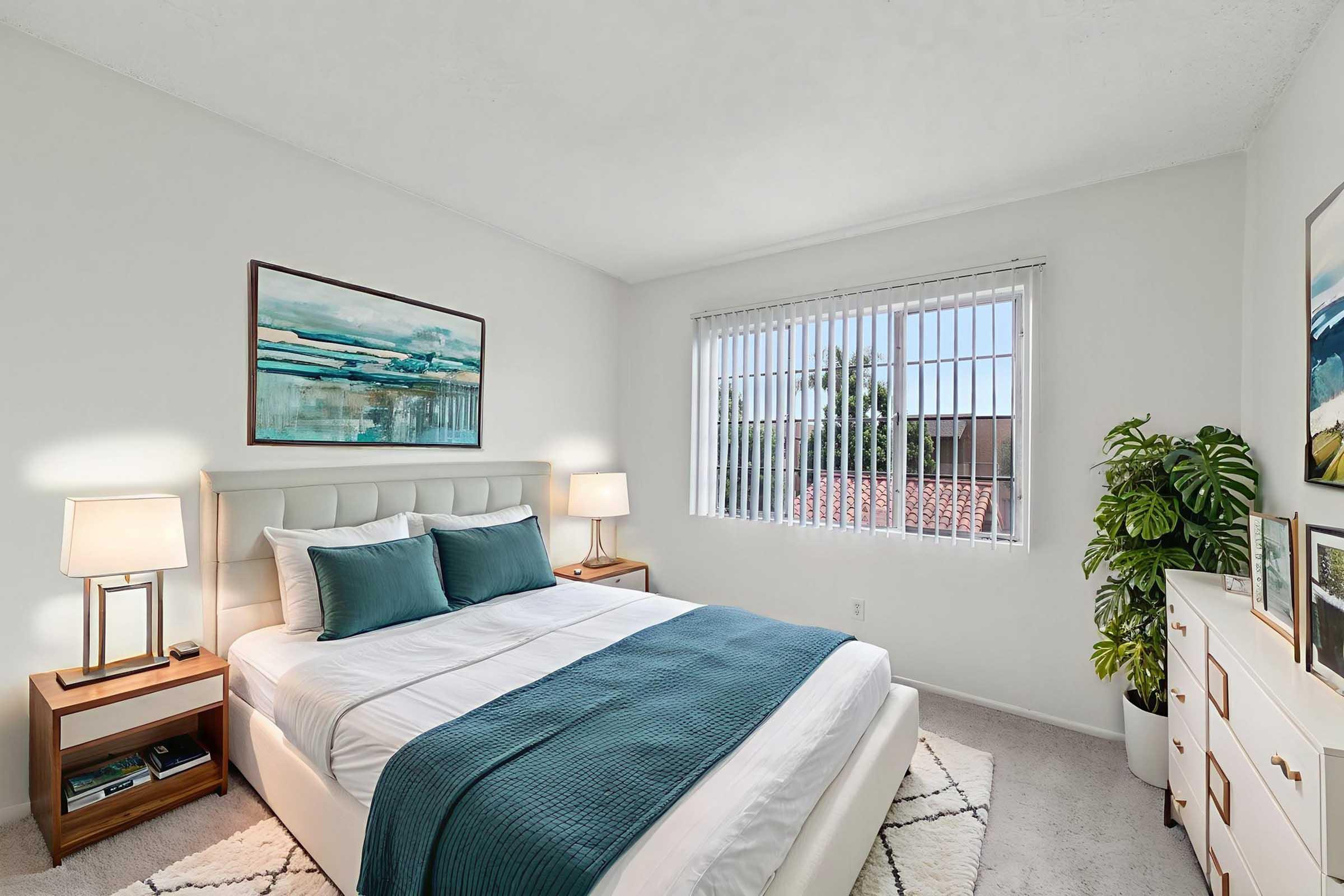 A bright and airy bedroom featuring a queen-size bed with a white and teal bedding set. Beside the bed are two wooden nightstands with lamps. A large window with horizontal blinds lets in natural light, and there’s a decorative plant in the corner. Wall art adds a modern touch to the minimalist decor.