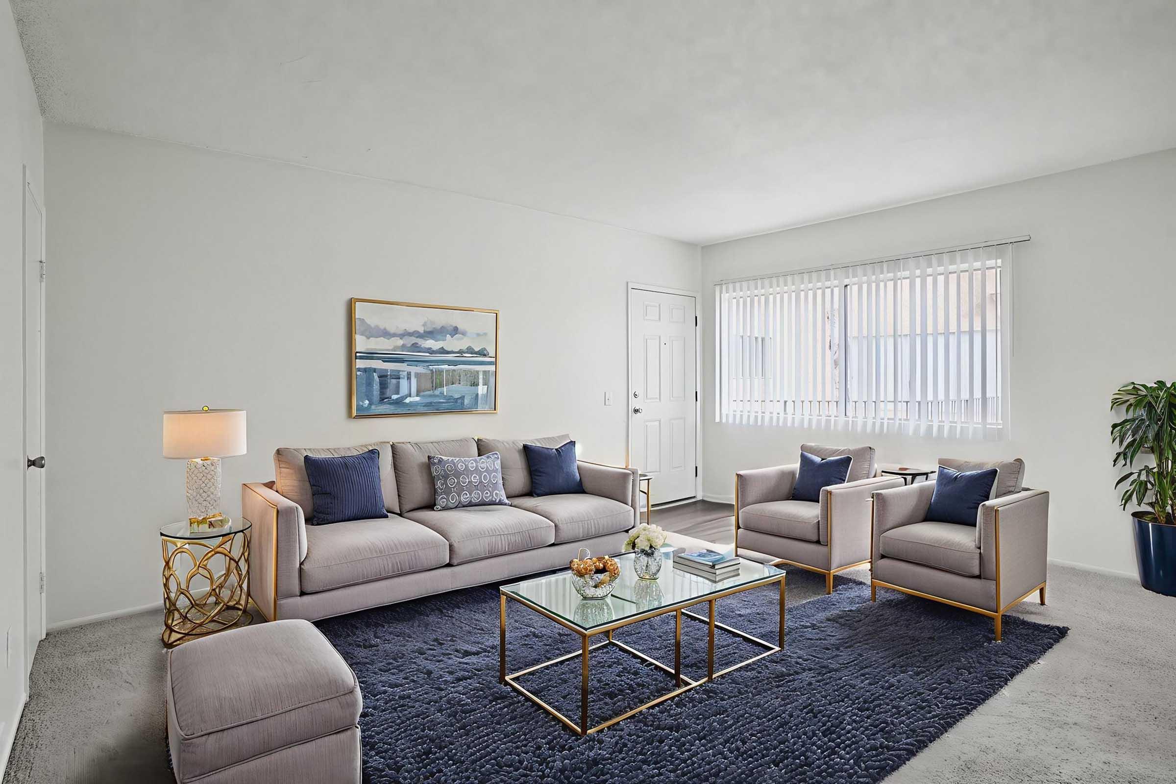 A modern living room featuring a light-colored sofa with blue pillows, two matching armchairs, and a coffee table. There is a decorative lamp beside the sofa, a piece of artwork on the wall, and a potted plant in the corner. Natural light comes through the window with blinds.