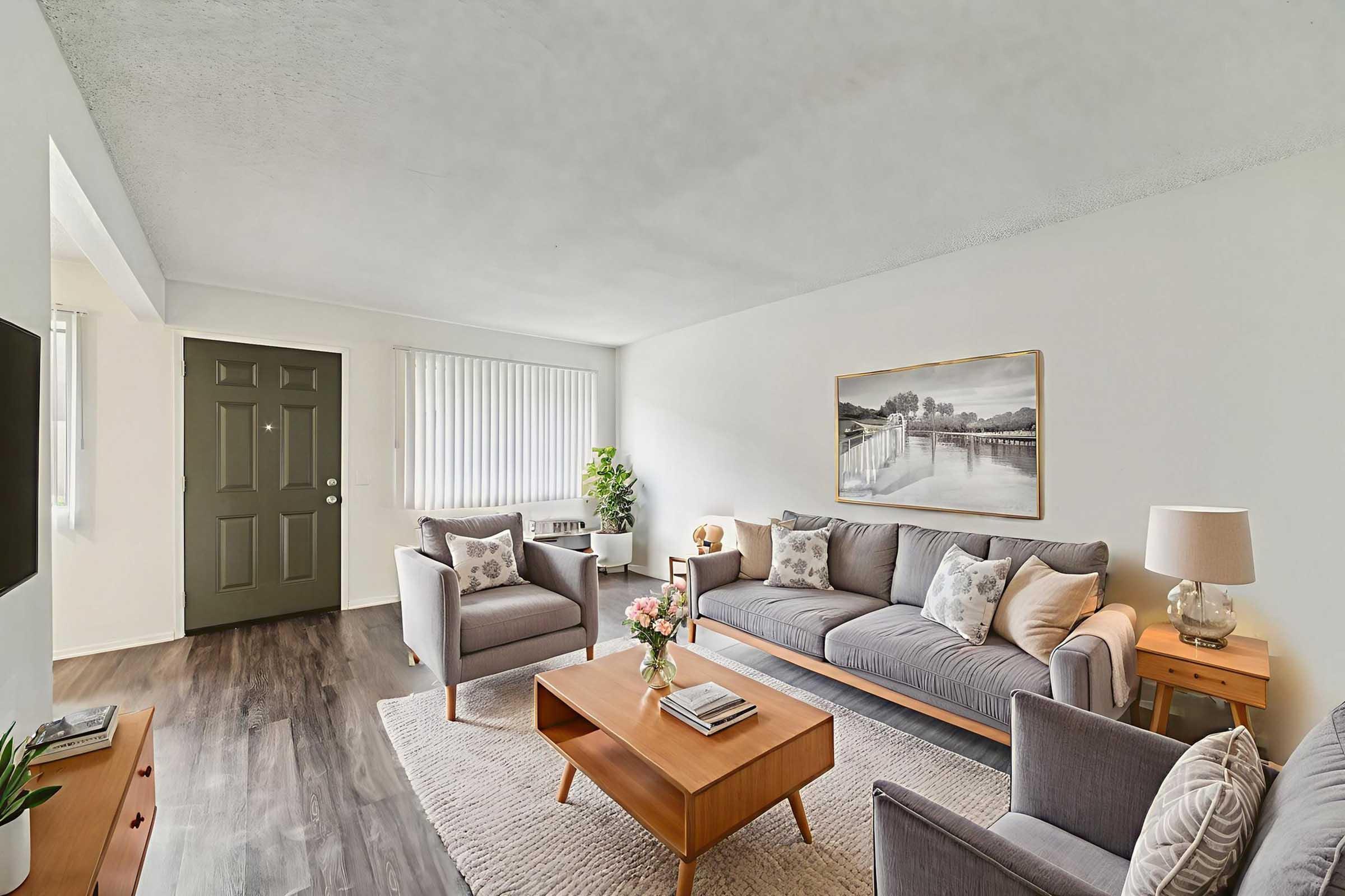 A cozy living room featuring a gray sofa and two matching armchairs, arranged around a wooden coffee table with flowers. A framed landscape painting hangs on the wall, with a door leading outside visible. Natural light filters through the window with vertical blinds, enhancing the inviting atmosphere.