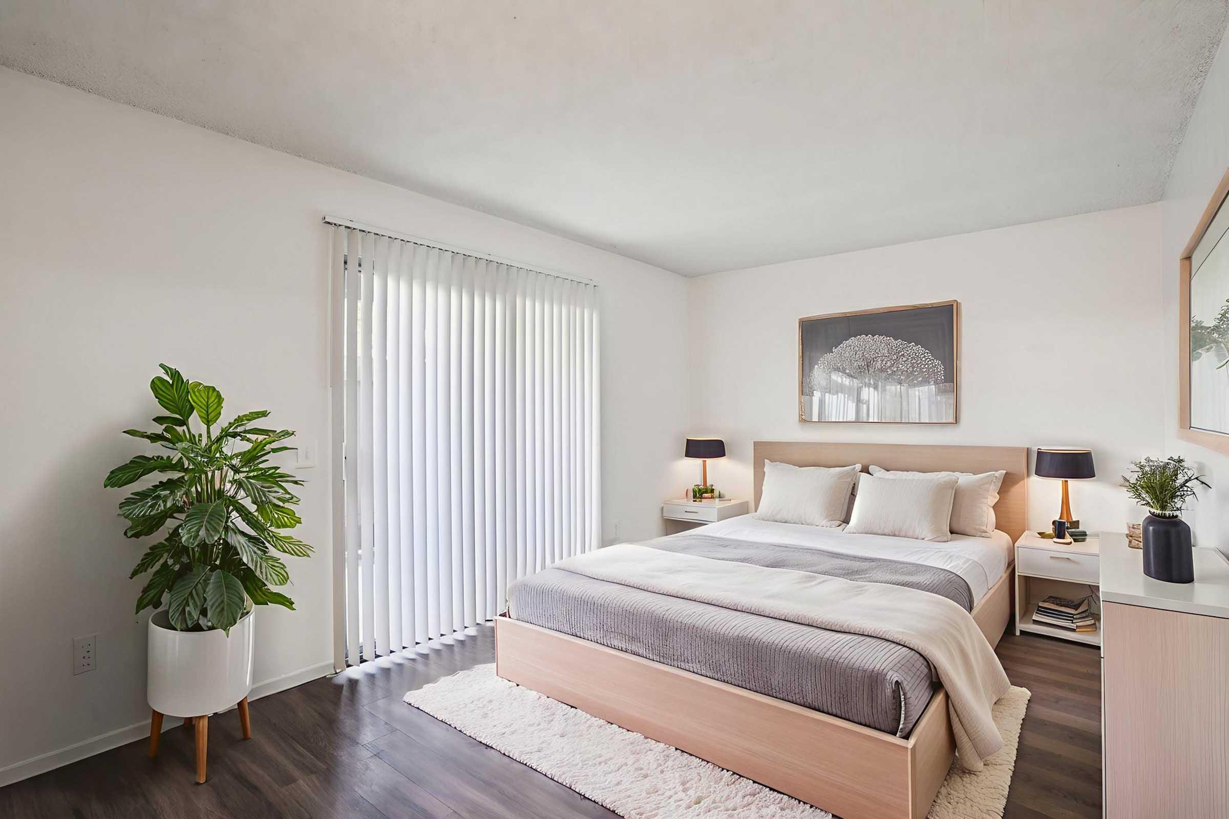 A cozy bedroom featuring a wooden bed with gray bedding, two bedside lamps, and a small nightstand. A large plant is placed in a white pot near the window, which has vertical blinds. A framed artwork hangs above the bed, complementing the neutral color palette and modern decor.