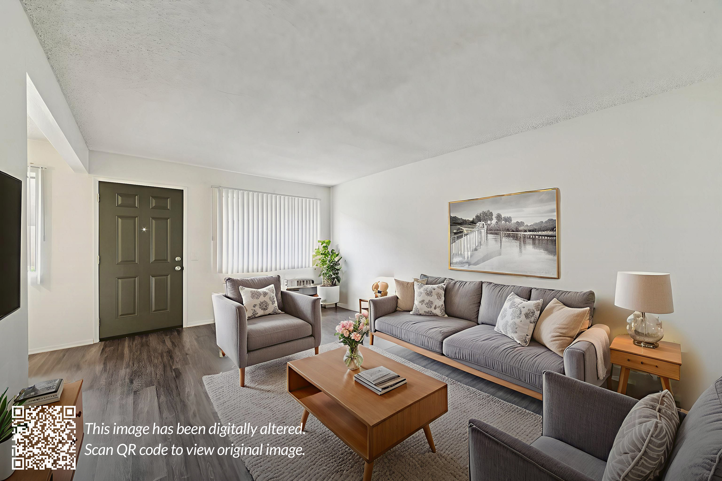 A modern living room featuring a light-colored sofa with decorative cushions, a wooden coffee table, and an artwork on the wall. There are gray armchairs and a potted plant, as well as large windows allowing natural light. The flooring is dark wood, adding warmth to the space.