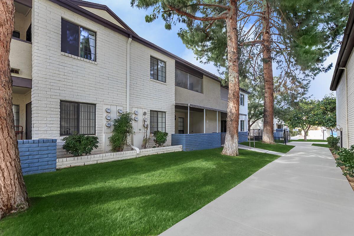 A view of a residential building with a landscaped yard featuring grass and trees. The building has several windows and a pathway leading alongside it.