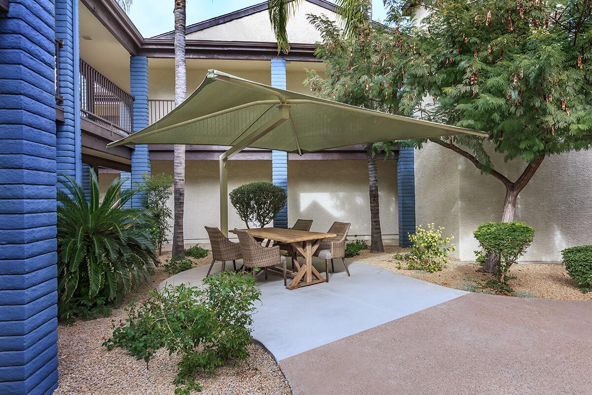 Outdoor seating area with a wooden table and four chairs under a large shade sail, surrounded by green shrubs and plants in a landscaped courtyard. The setting features neutral-colored walls and a blue column, with palm trees in the background.