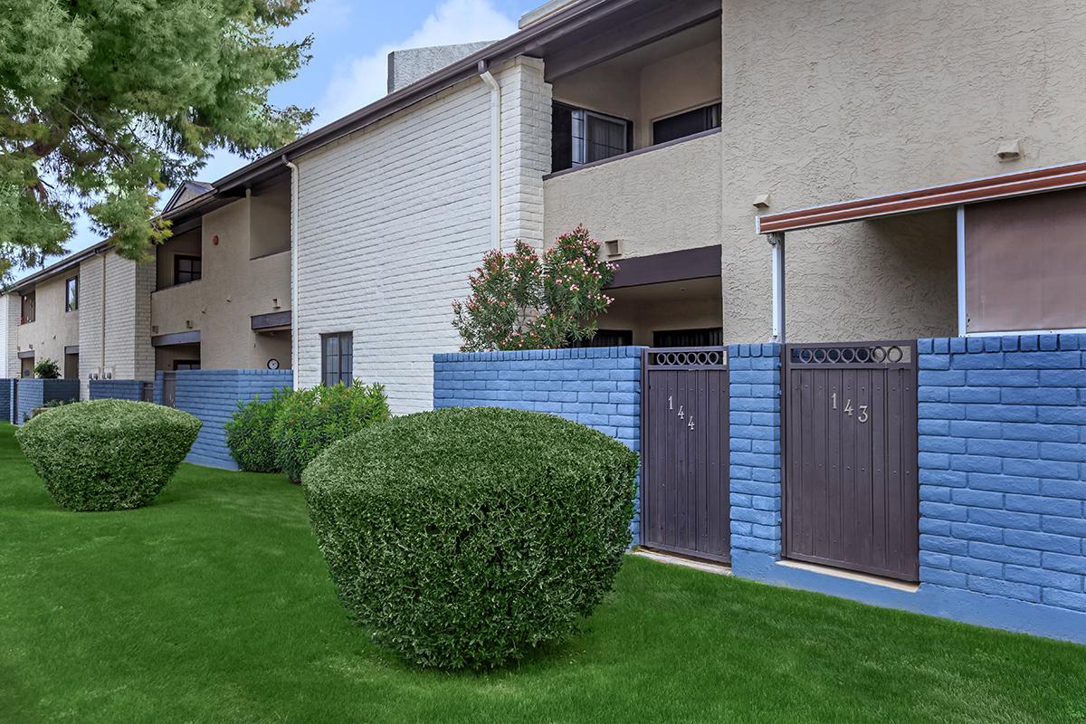 A row of apartment buildings with a neatly manicured lawn, featuring green bushes in front of blue and brown brick walls. Each apartment has a door with the numbers 143 prominently displayed.