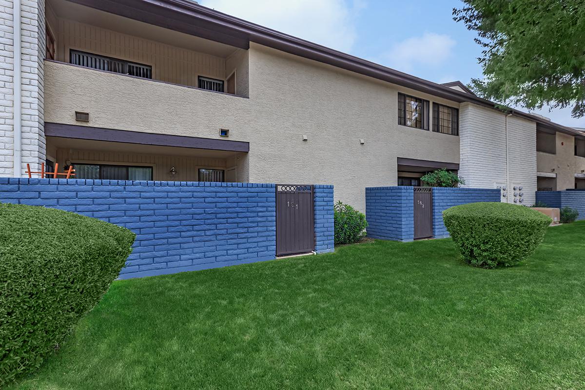 A view of a residential building with two stories, featuring a light-colored exterior and blue painted walls around the entrances. There are manicured green lawns and some bushes in front of the building. The sky is partly cloudy.