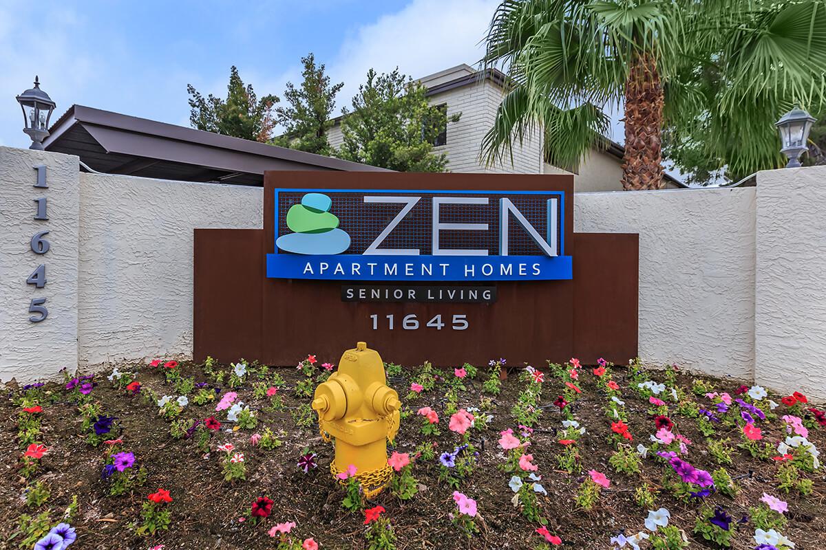 Sign for Zen Apartment Homes, featuring the name in blue and green lettering, with the subtitle "Senior Living." The sign is surrounded by landscaped flower beds and includes a yellow fire hydrant.