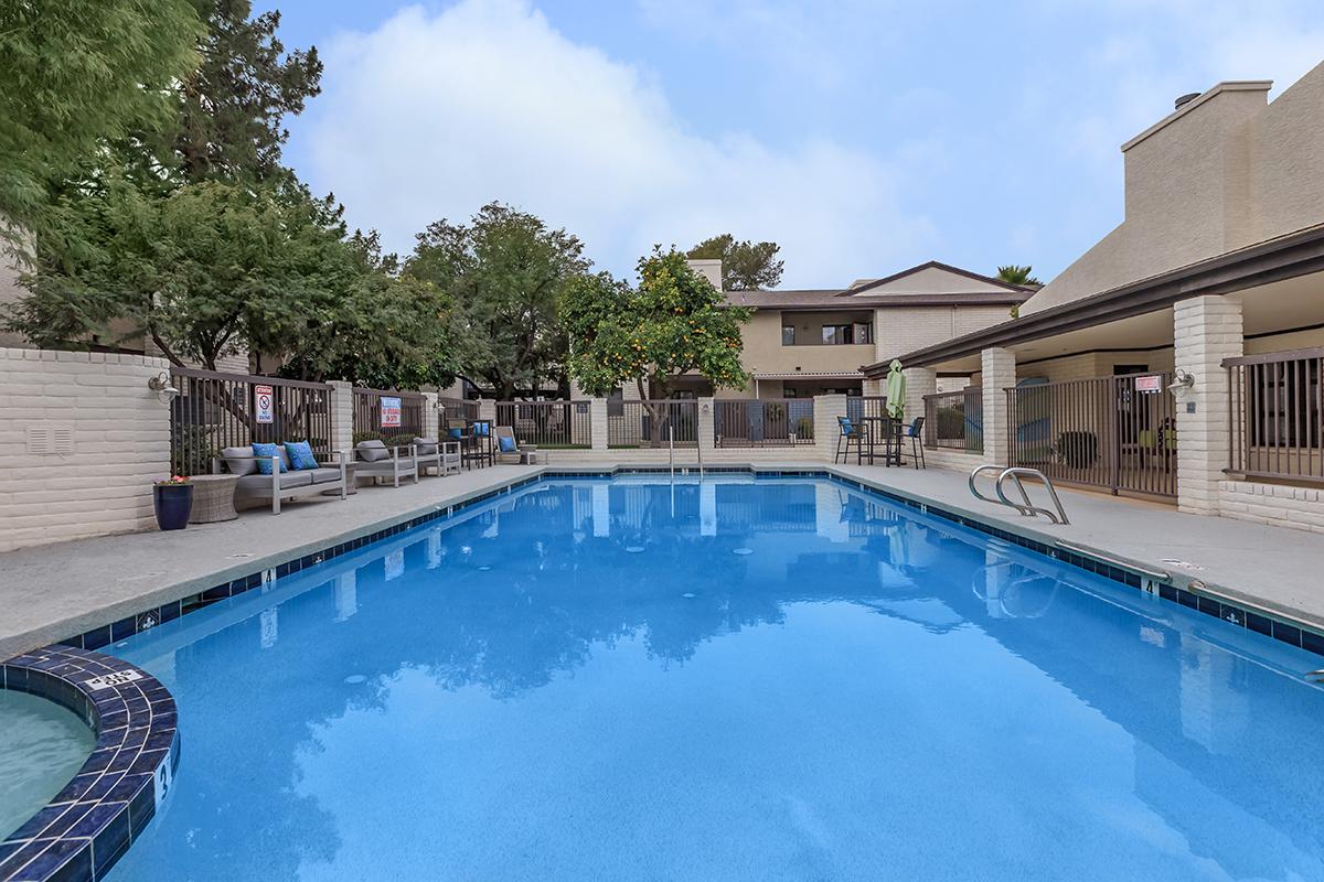 A clear blue swimming pool surrounded by lounge chairs and patio furniture, with trees and buildings in the background under a partly cloudy sky.