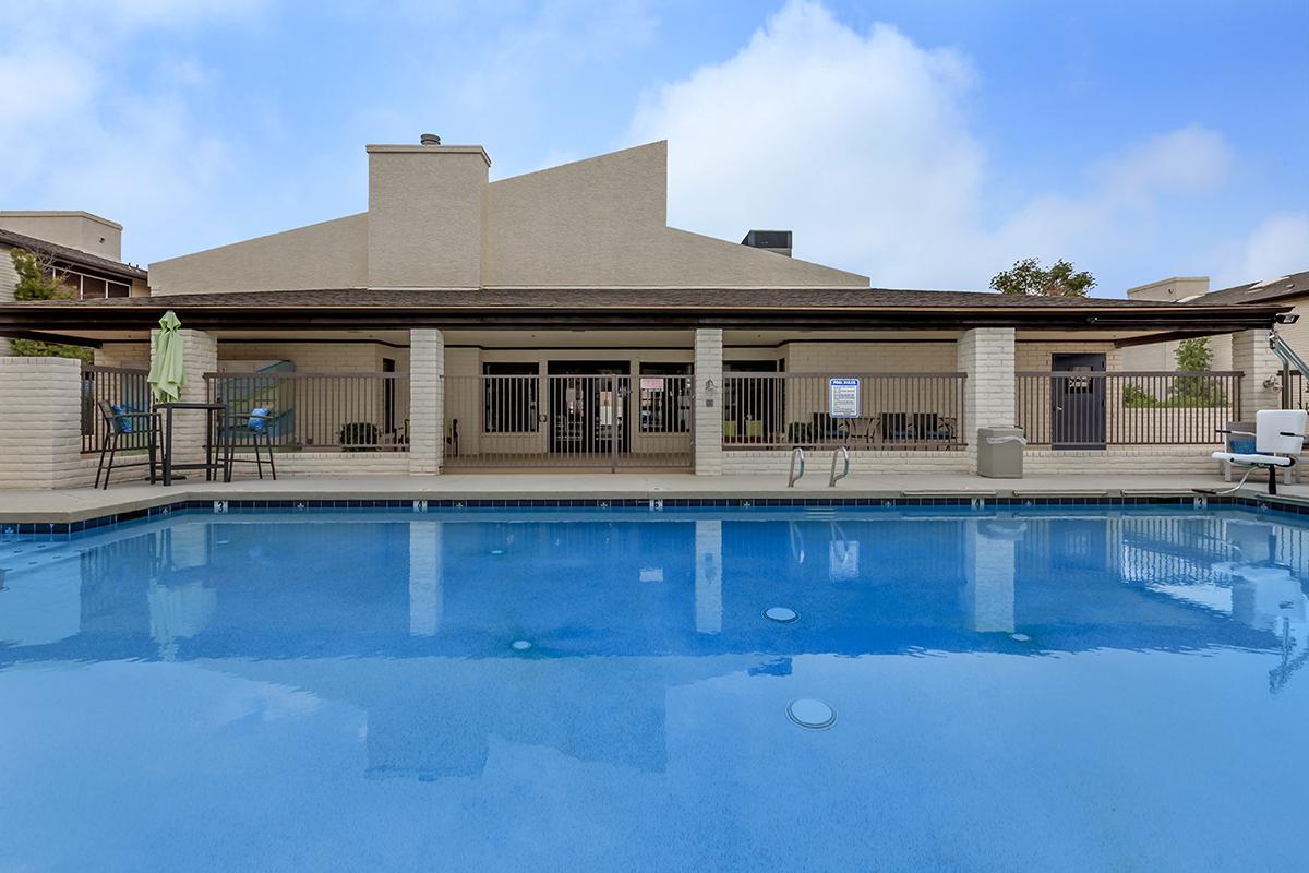 View of a swimming pool with clear blue water, surrounded by a patio area featuring lounge chairs and a shaded seating section. In the background, there is a building with a flat roof and a fenced area. The sky is partly cloudy.