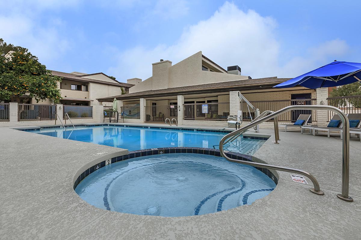 A swimming pool area featuring a blue pool with a hot tub integrated into the design. Surrounding the pool are lounge chairs and an umbrella. In the background, there are buildings and a fence, with a clear sky above.