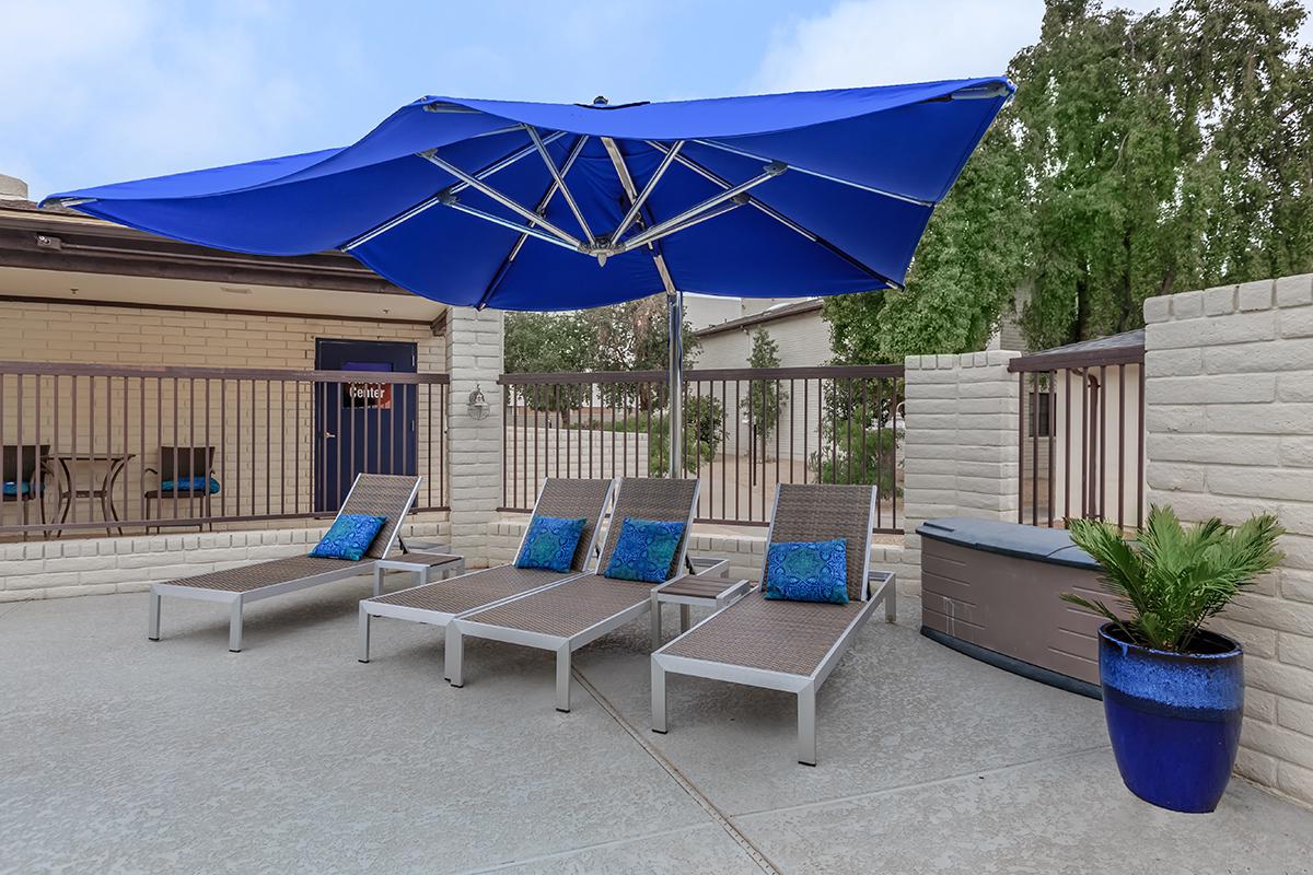 A poolside area featuring four lounge chairs with blue cushions positioned under a large blue umbrella. There is a potted plant in a blue planter and a hot tub visible nearby, set against a backdrop of a light-colored wall and trees.