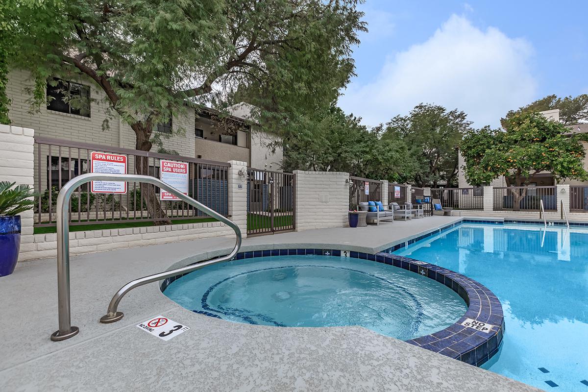 A pool area featuring a spa with a railing and a swimming pool, surrounded by greenery and residential buildings. Safety signs are posted around the area, and lounge chairs are visible near the pool. The sky is partly cloudy.