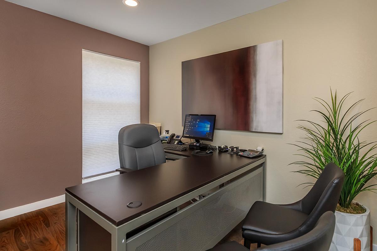 Office interior featuring a modern desk with a computer, phone, and office supplies. A comfortable black chair is positioned at the desk, with two additional black chairs nearby. A large abstract painting hangs on the wall, and a potted plant adds a touch of greenery. Natural light filters through a window with shades.