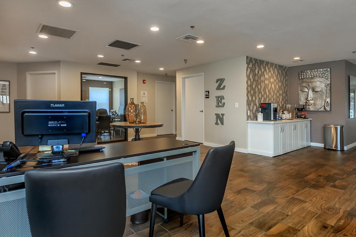 A modern office reception area with a desk featuring a computer, black chairs, and decorative elements. The walls are painted in neutral tones with a Buddha sculpture and the word "ZEN" displayed. The flooring is wooden, and there are plants and glass vases as decor.