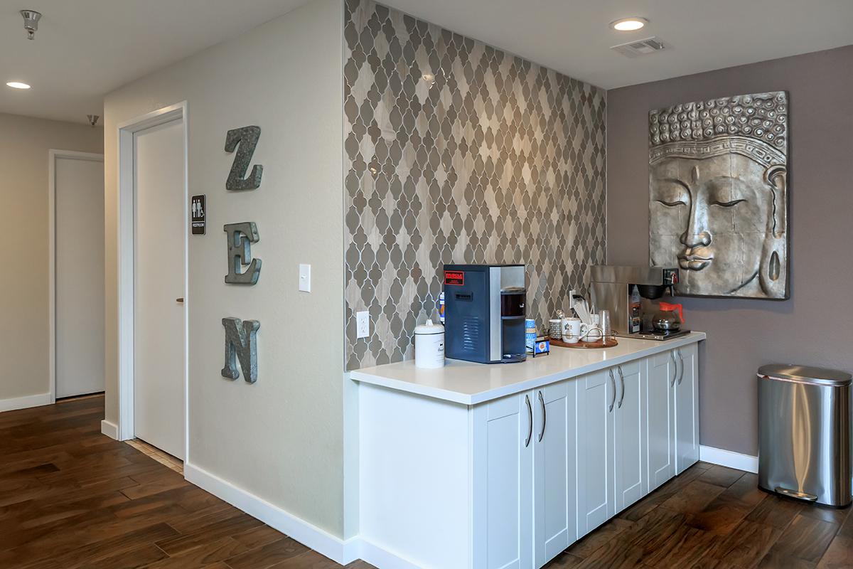 Modern office kitchenette with a coffee machine, cabinets, and a decorative Buddha wall art. The wall features a textured pattern, and the word "ZEN" is displayed prominently. Wood flooring and a trash bin are visible in the space.