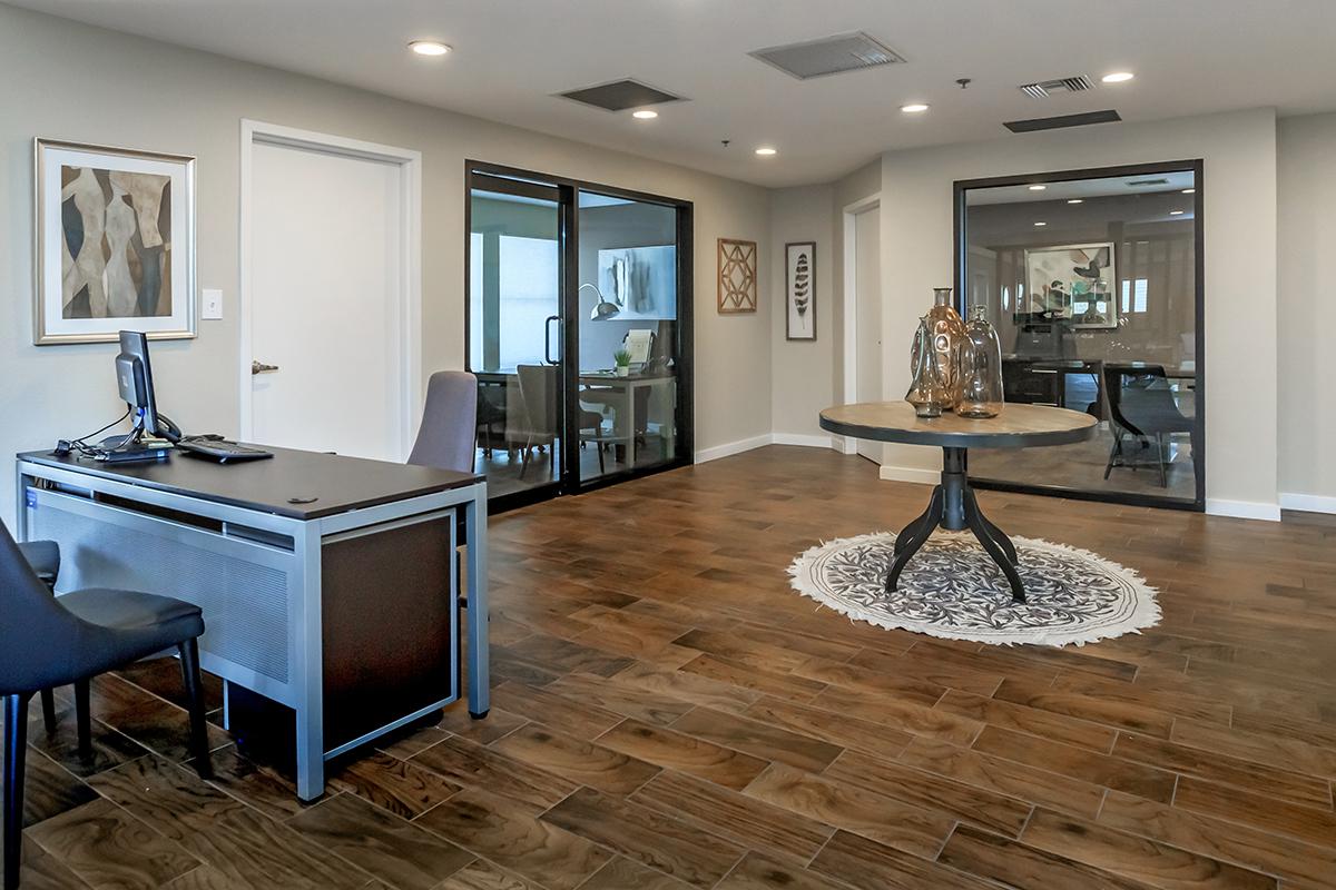 A well-lit interior space featuring a modern office setup with a desk and chair on the left. In the center, there is a round wooden table with decorative vases on top, surrounded by a patterned rug. The background includes glass doors leading to another room, creating an open, inviting atmosphere.