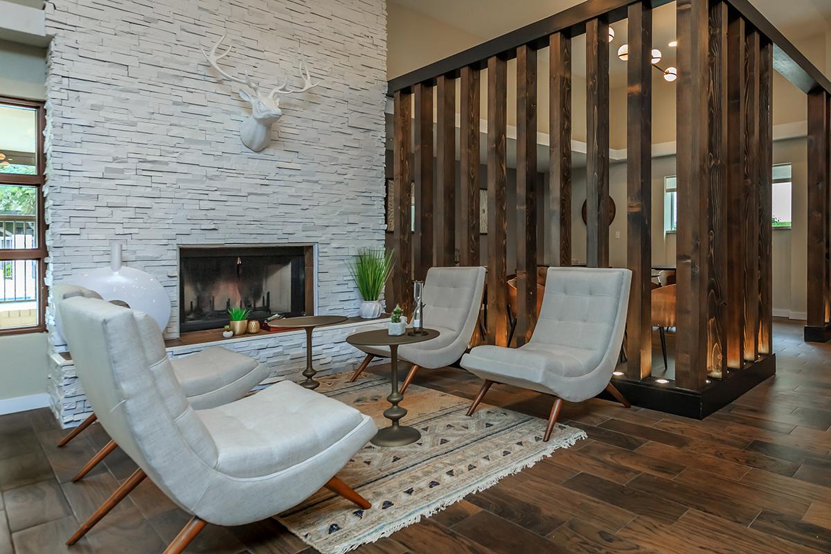 A modern living space featuring four light gray chairs arranged around a small round table, with a stone fireplace and a decorative deer head mounted on a textured wall. Wooden slatted partitions create separate areas, and a patterned rug adds warmth to the wooden flooring.